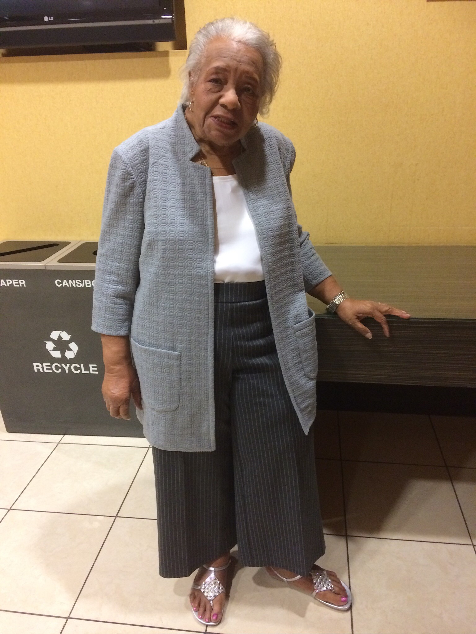 Older woman with gray hair wearing a light blue textured blazer, white blouse, pinstriped pants, and silver sandals, standing by a recycling bin in a indoor setting