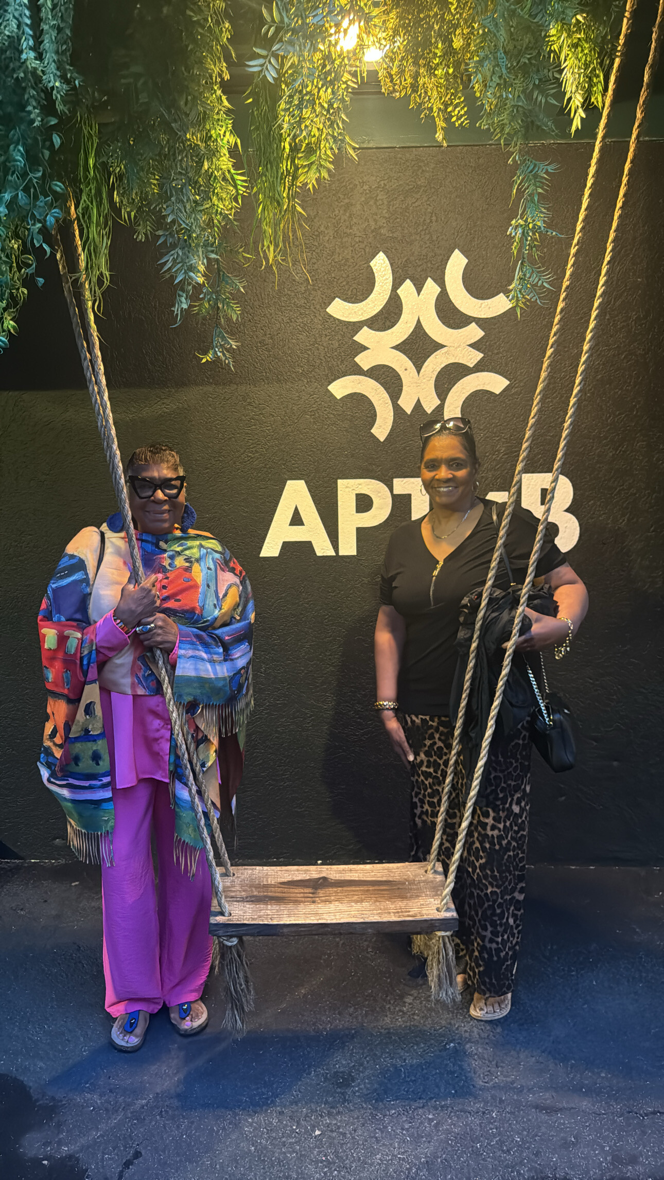 Two smiling women stand on a rope swing against a dark wall with a white geometric logo and hanging greenery above.