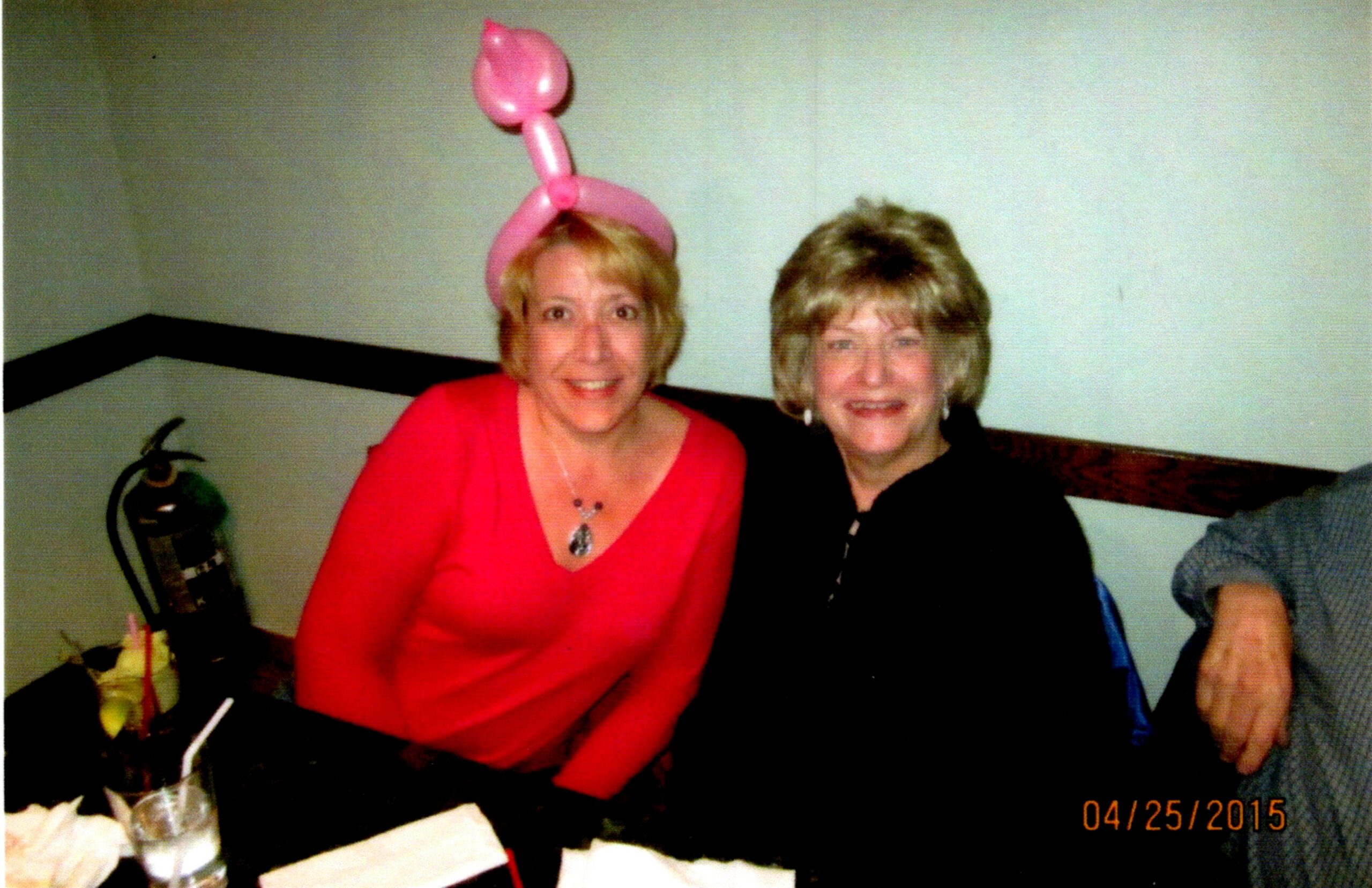 Two smiling women sit at a party table; the left woman wears a pink balloon hat and red top.