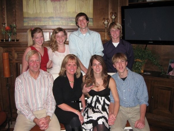 Eight people pose for a two-row group photo in a living room, smiling and dressed casually for a gathering.
