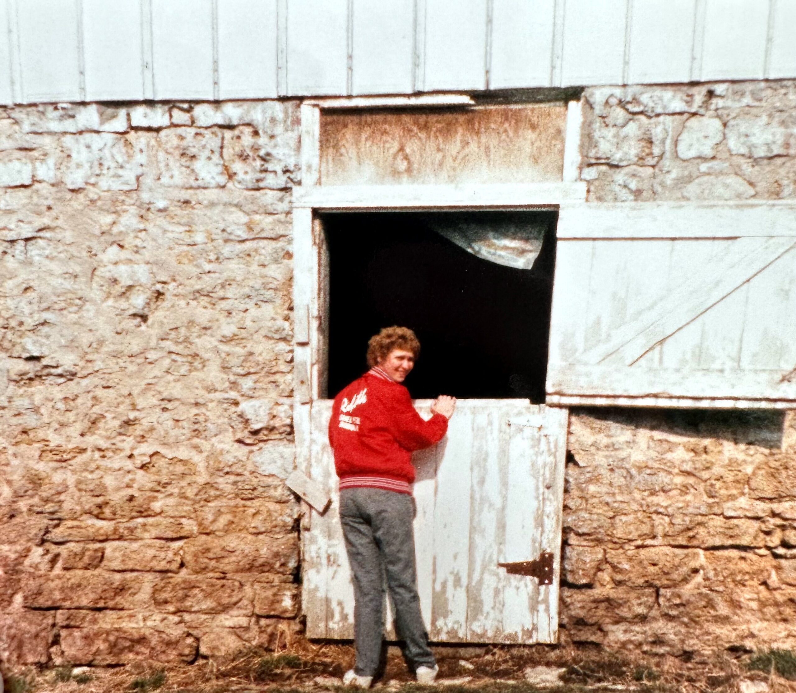 Man in a red jacket standing on a small wooden balcony/ledge outside a rough stone building, looking toward the camera.