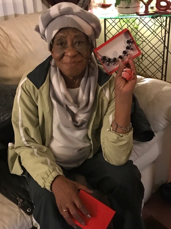 Older woman sitting on a couch, wearing a head wrap and scarf, showing a red jewelry box with a beaded necklace.