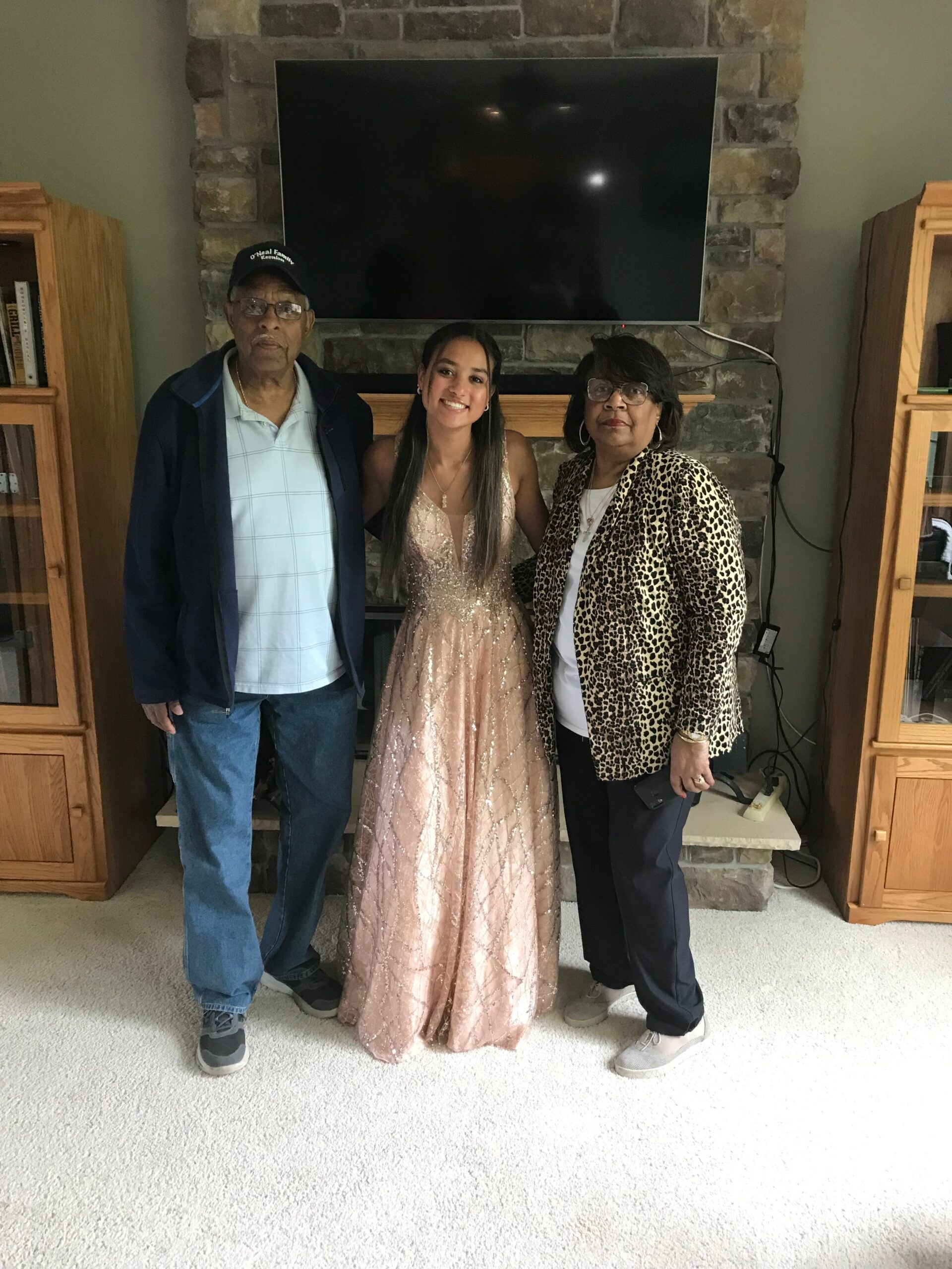 Three people pose together in a living room in front of a stone fireplace and flat-screen TV above. The young woman in a pink, sparkly gown is flanked by an older man in a cap and a woman in a leopard-print jacket.