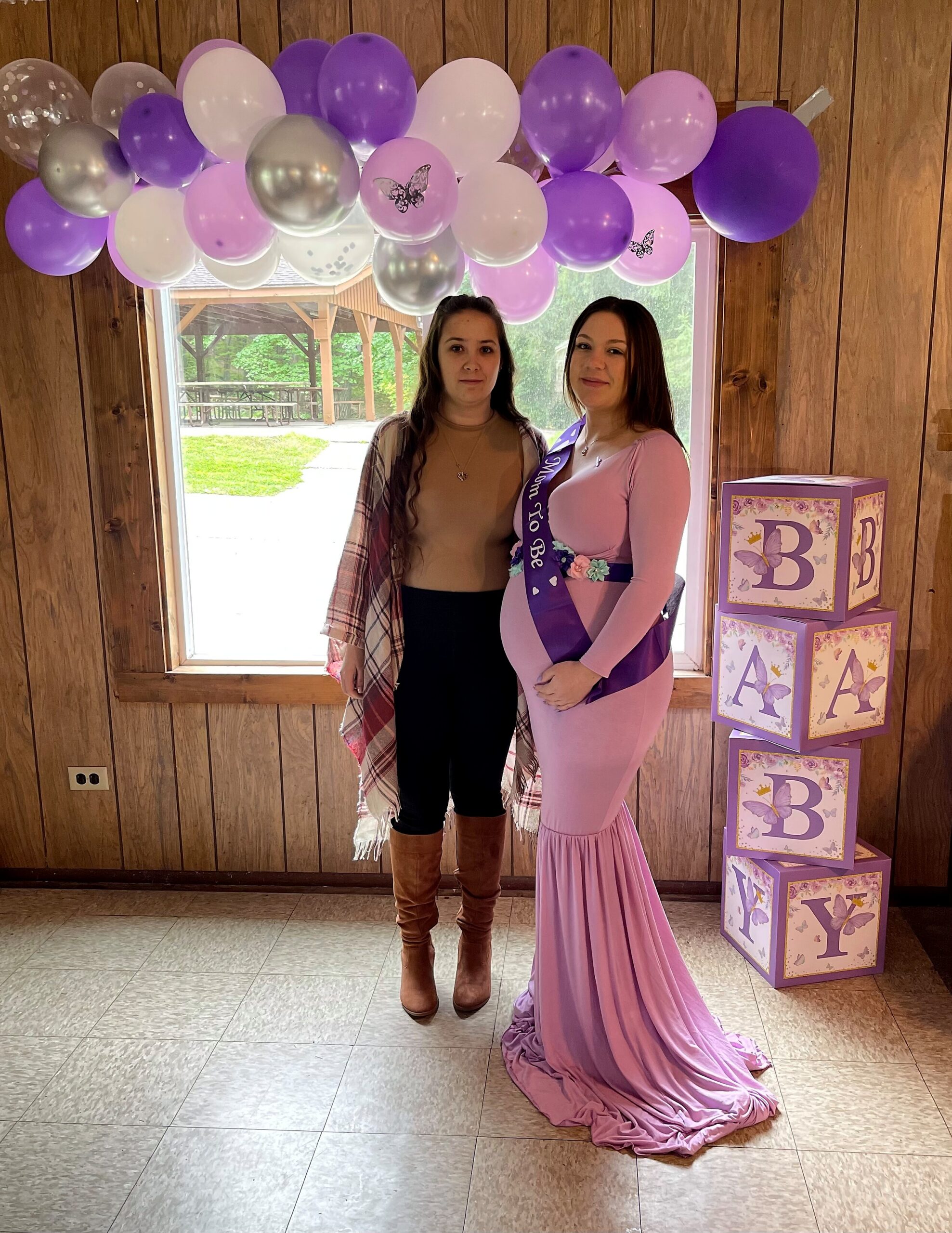 Two women pose indoors under a purple and pink balloon arch; one wears a pink maternity dress with a 'Mom To Be' sash, the other in a plaid shawl.