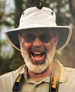 Man wearing a wide-brim beige hat and sunglasses, laughing outdoors.