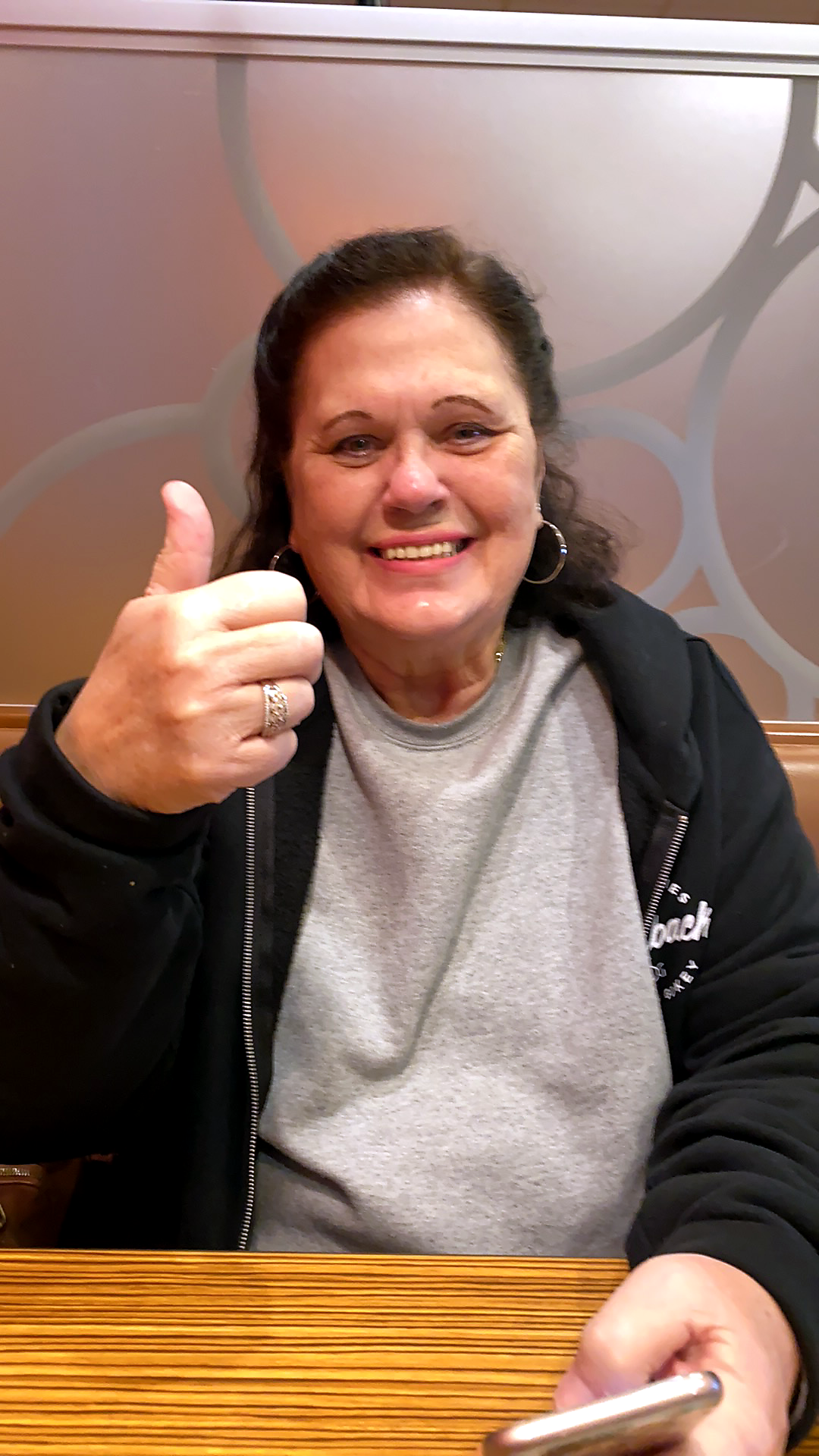 Smiling woman giving a thumbs-up, seated at a wooden table, wearing a gray shirt and black hoodie/ jacket behind.