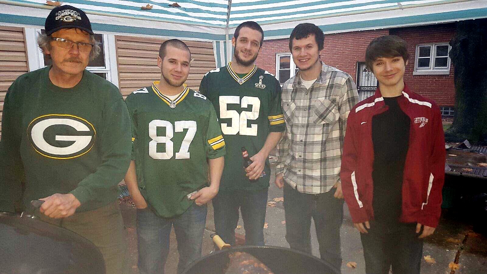 Five men pose together in a driveway under a striped canopy; two wear Green Bay jerseys, one in a plaid shirt, and one in a red jacket.