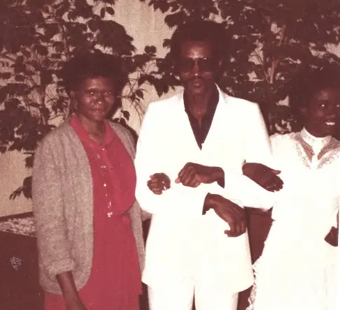 Three people pose indoors: a man in a white suit with sunglasses center, a woman in a red dress and cardigan left, and a person in white on the right.