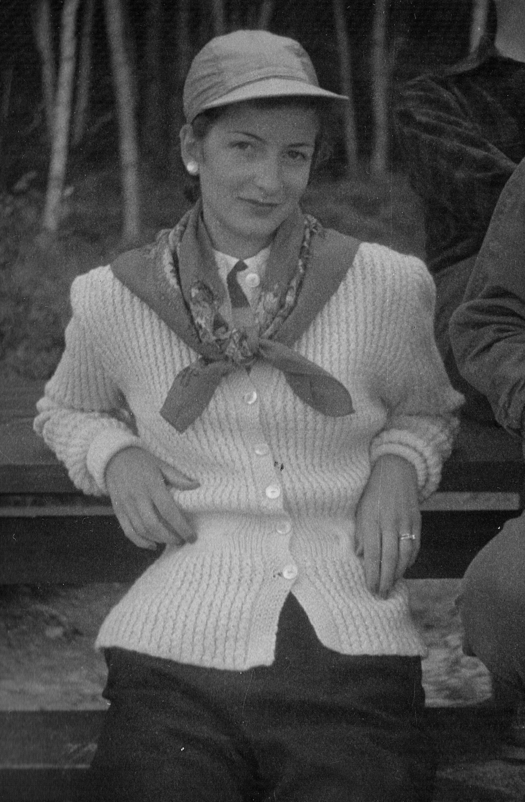 Black-and-white photo of a woman wearing a knit cardigan, scarf, and hat, seated and looking at the camera with a subtle smile.