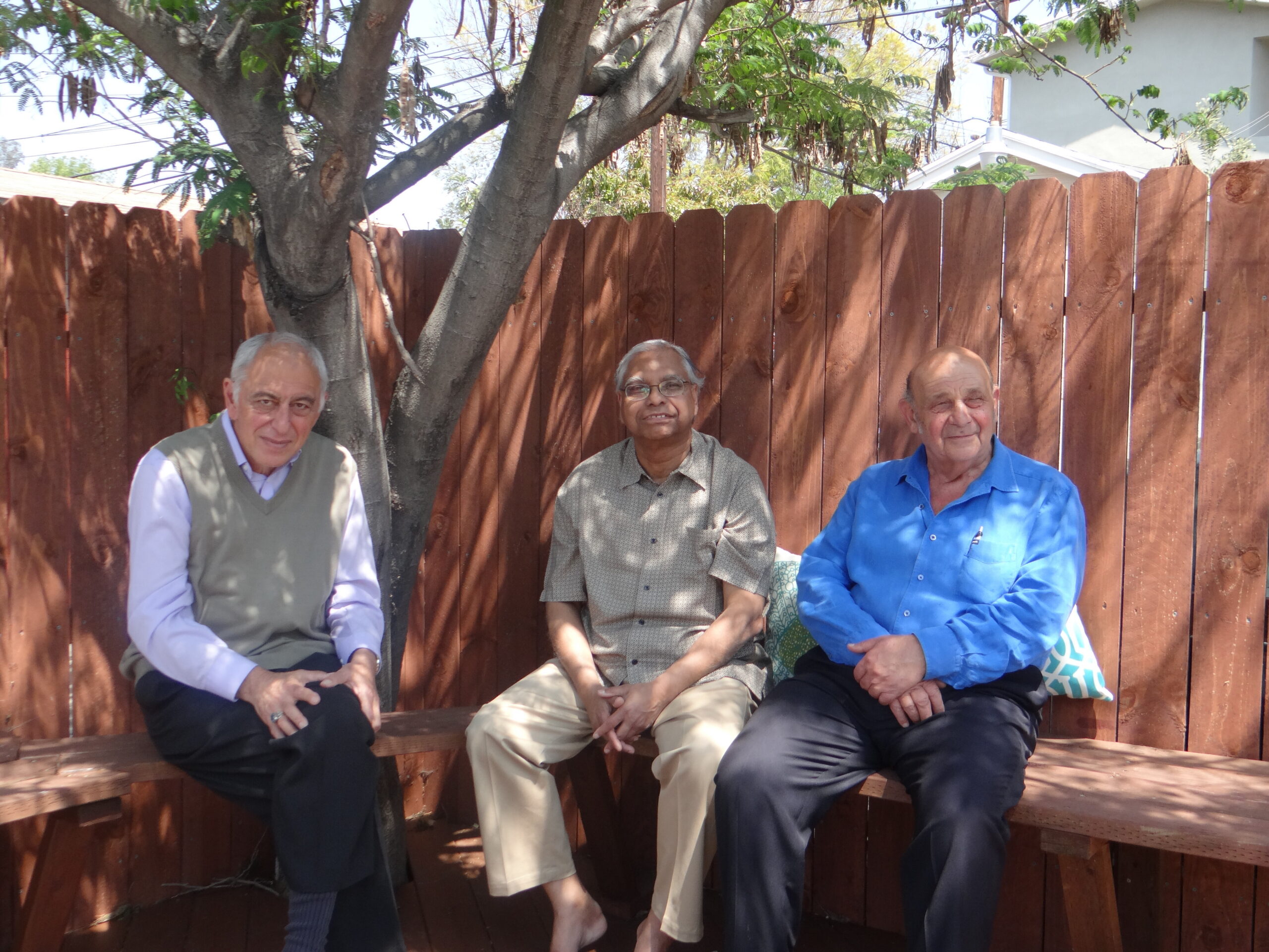 Three elderly men sit on a wooden bench in front of a tall wooden fence, smiling outdoors with a tree behind them.