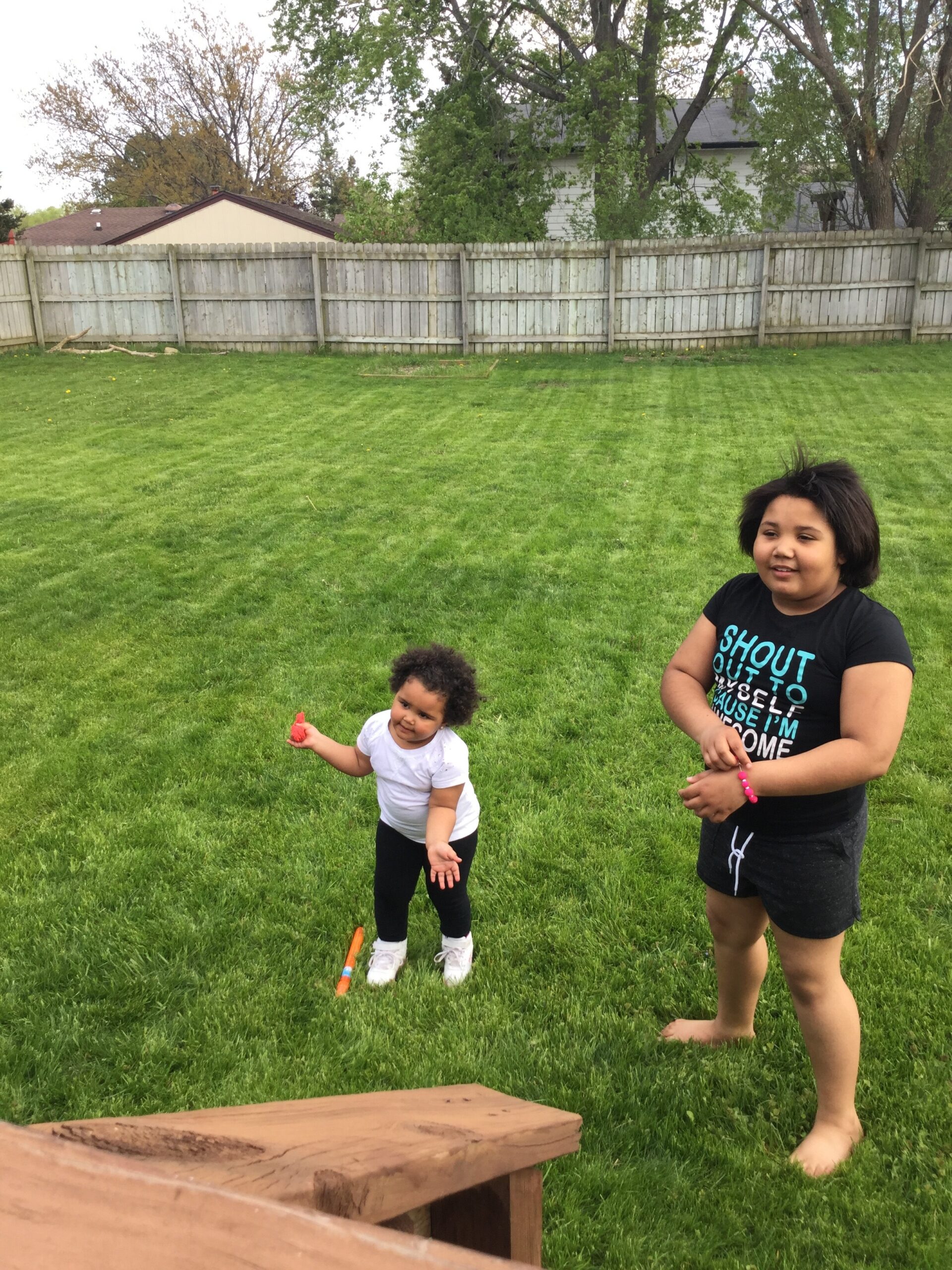 Two children playing on a green backyard lawn near a wooden bench and fence, one holding a small red toy.