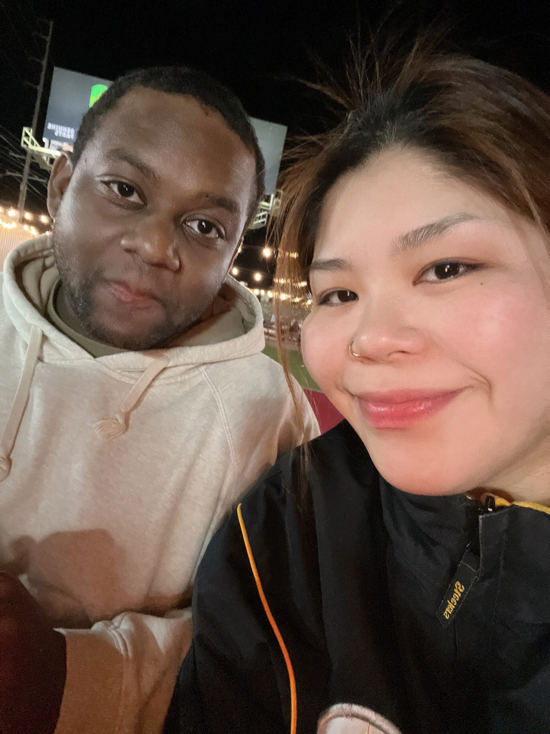 Two friends posing for a close-up nighttime selfie with string lights in the background. On the left, a man in a beige hoodie; on the right, a smiling woman with a nose ring.
