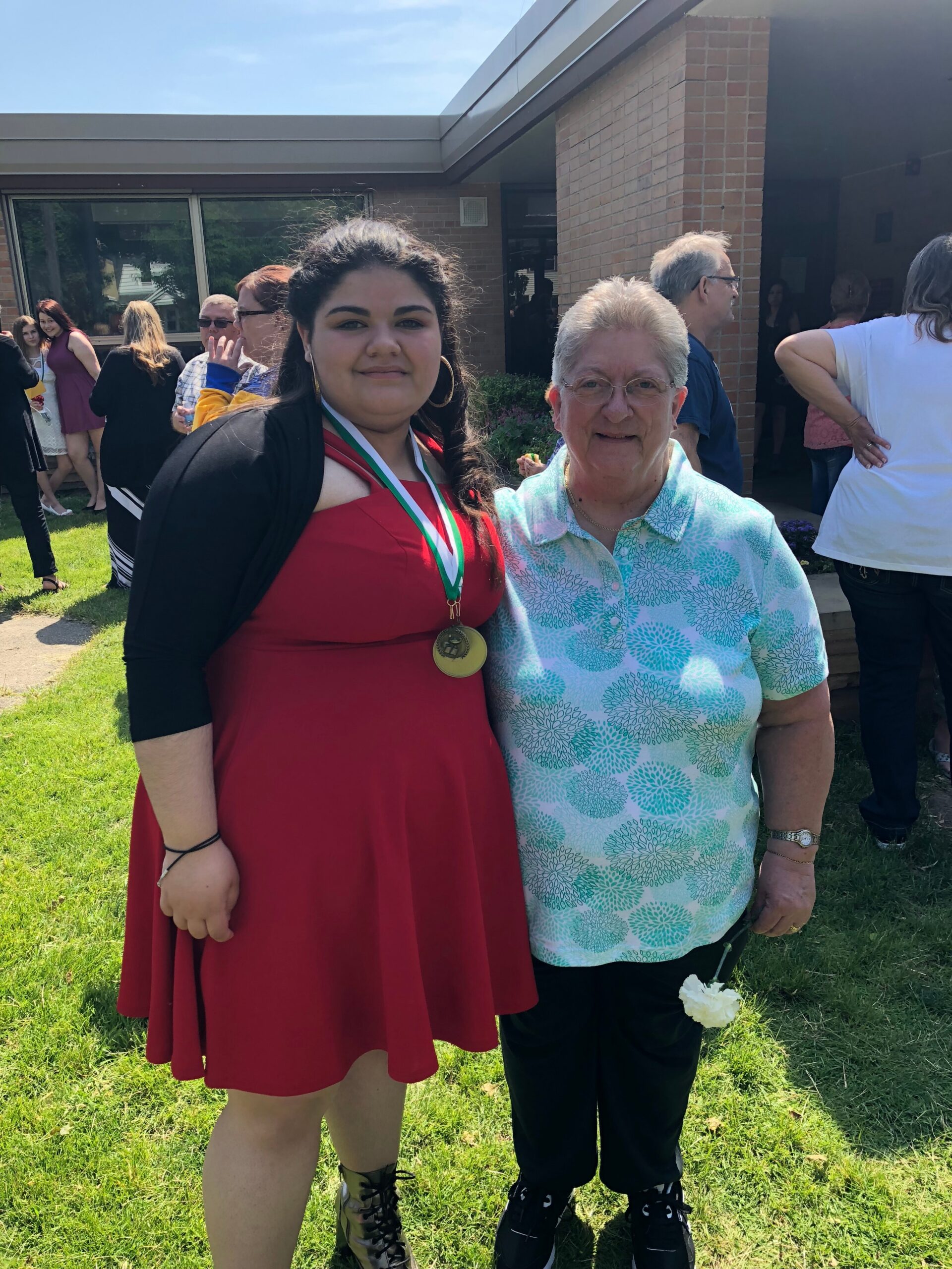 Young woman in a red dress with a gold medal around her neck stands beside an older woman in a teal floral shirt, outdoors near a brick building with a crowd in the background.