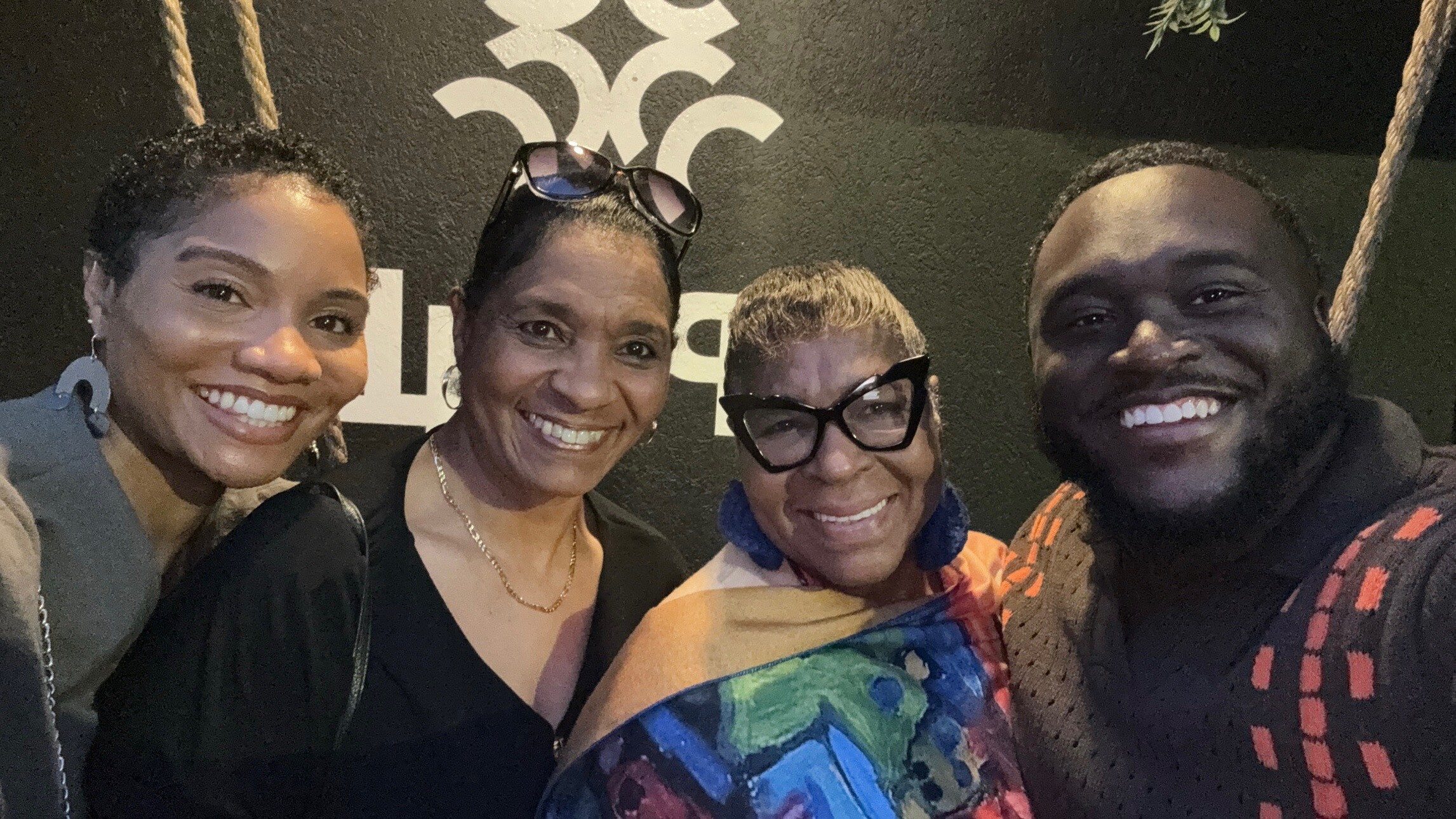 Four friends posing for a close-up group selfie, smiling at the camera indoors against a dark wall art background, with three women and one man.