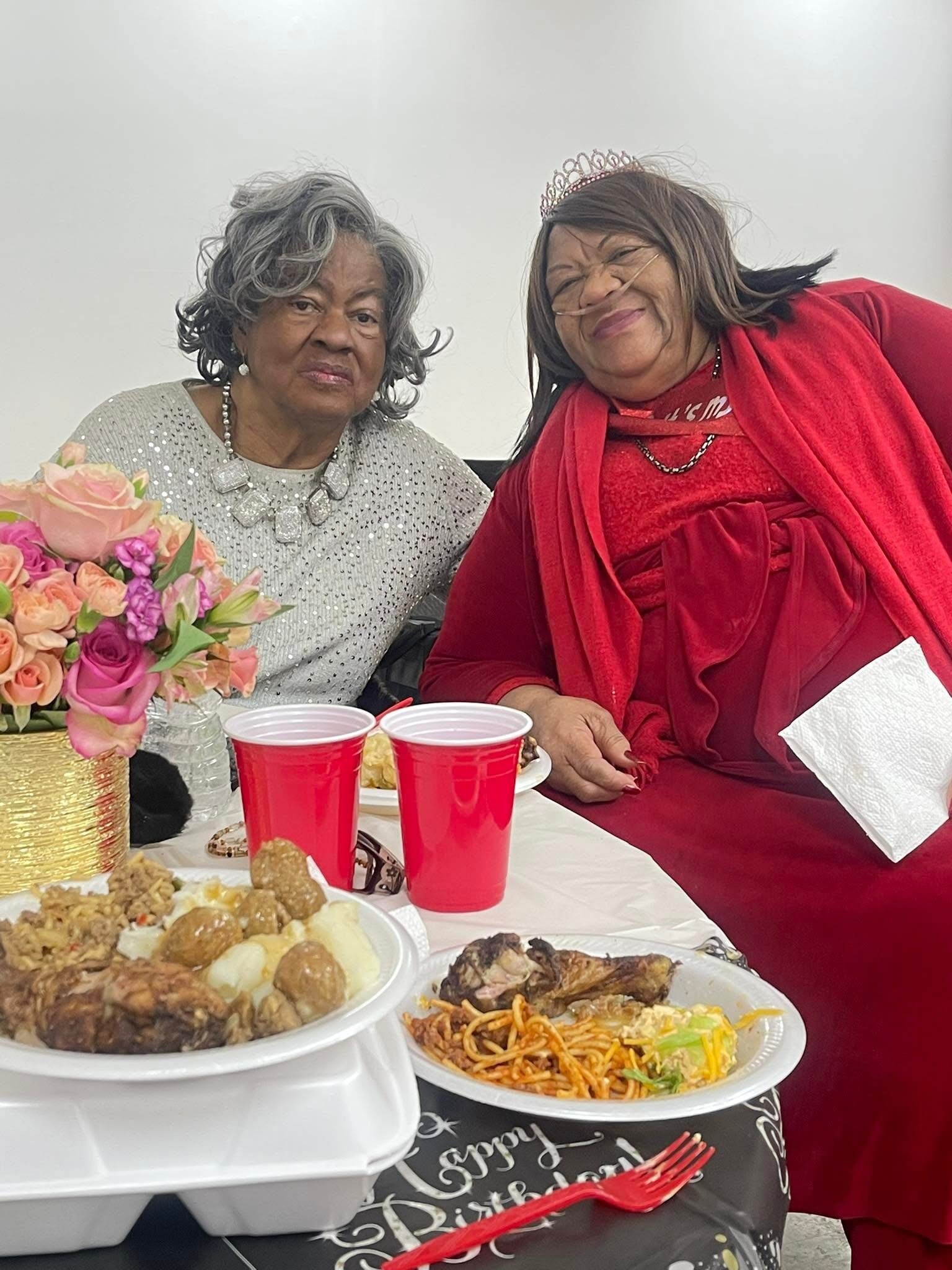 Two smiling women at a celebration, one in a silver sequined top with a bold necklace and the other in red with a tiara, seated by flowers and plates of food.