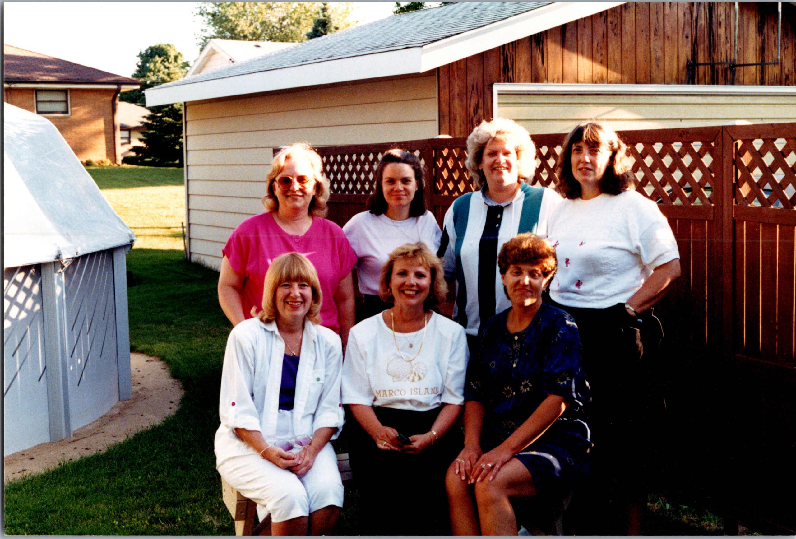 Group of women posing together in a backyard, with a lattice fence and houses in the background.