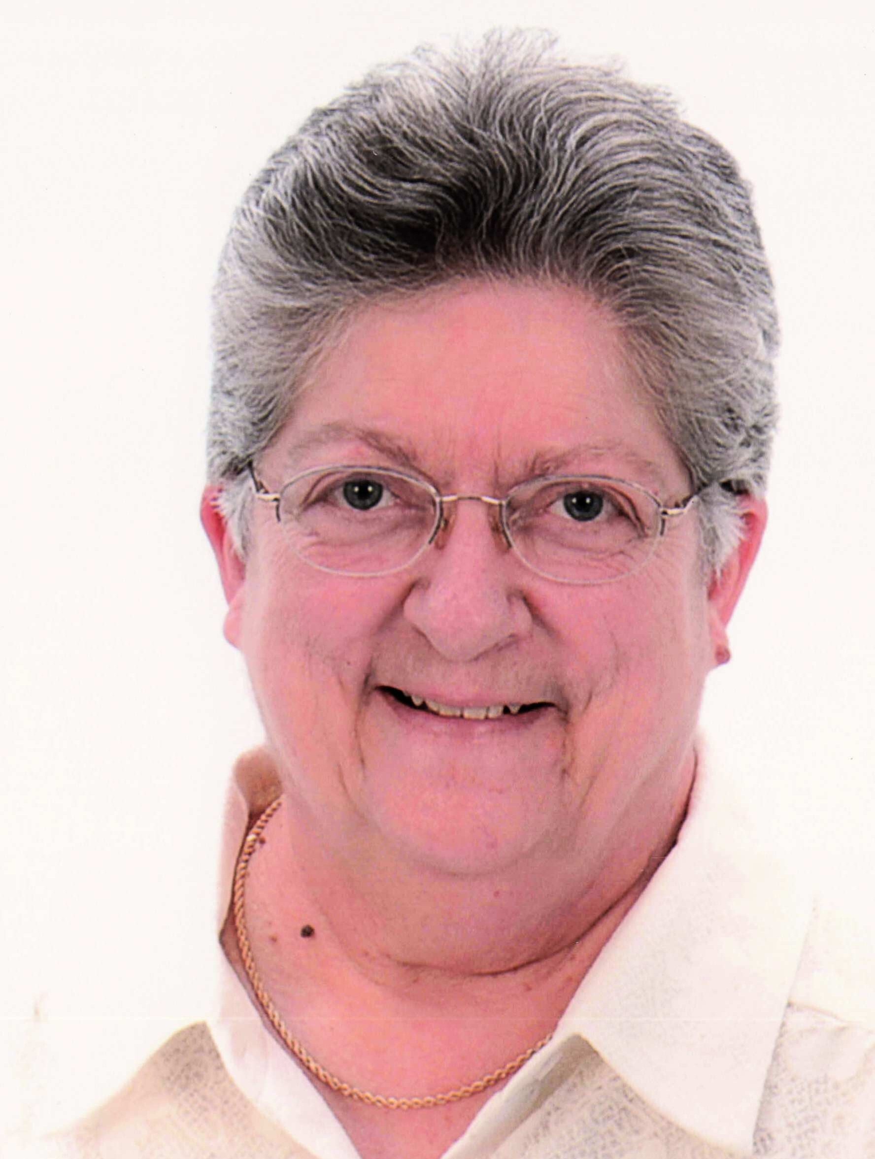 Portrait of an older woman with short gray hair and glasses, smiling gently against a light background, wearing a white collar and sweater vest with a gold chain.