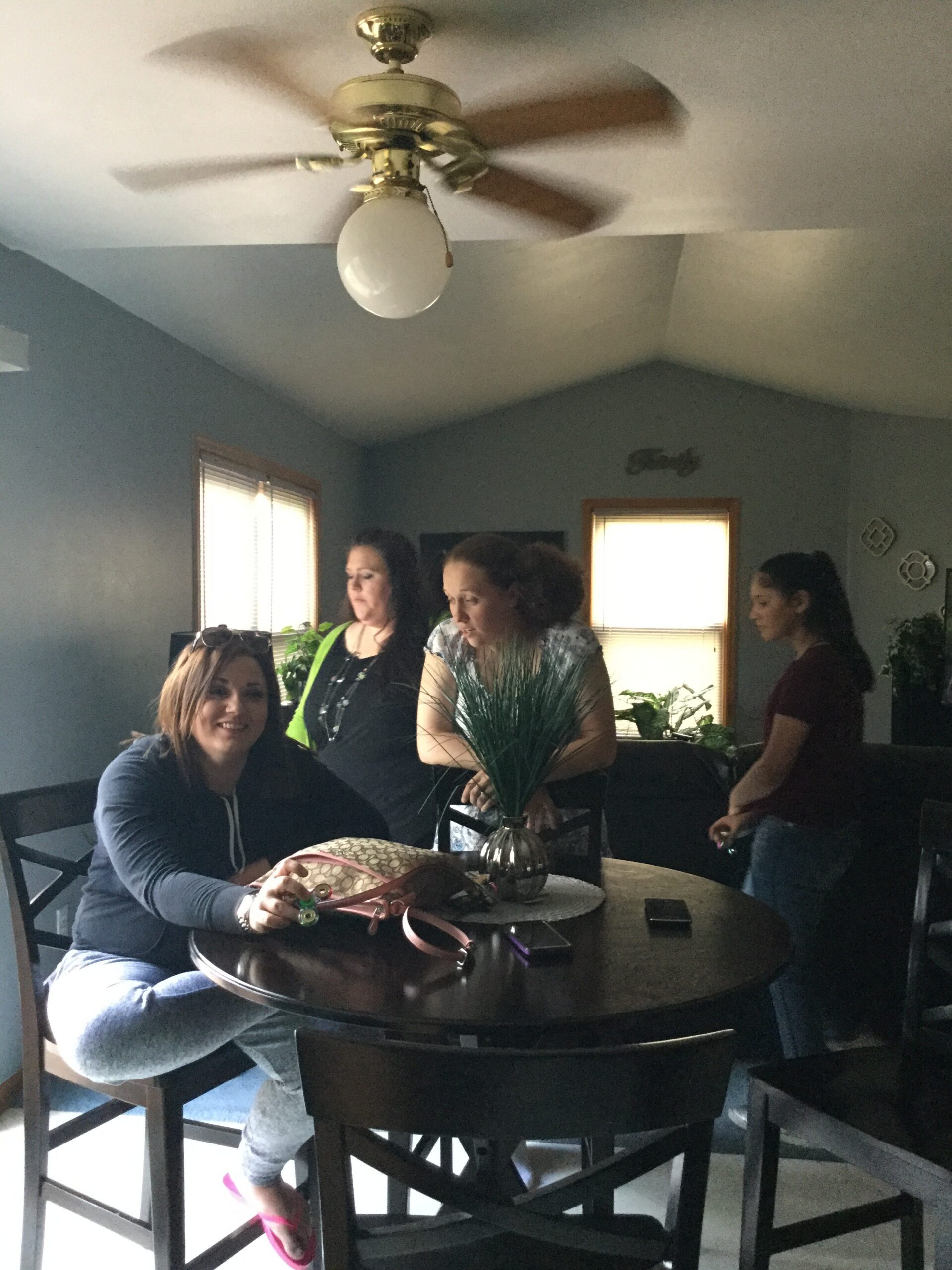 People gathered around a round dark dining table in a living room, with a ceiling fan overhead and windows letting in light.