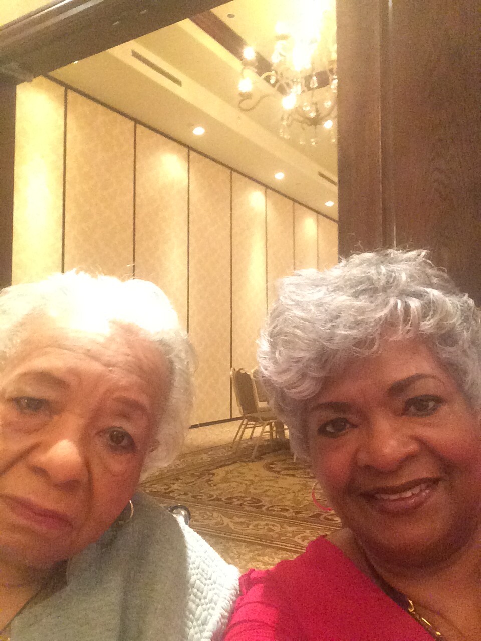 Two smiling women take a selfie in a chandelier-lit banquet hall with beige paneled walls behind them.