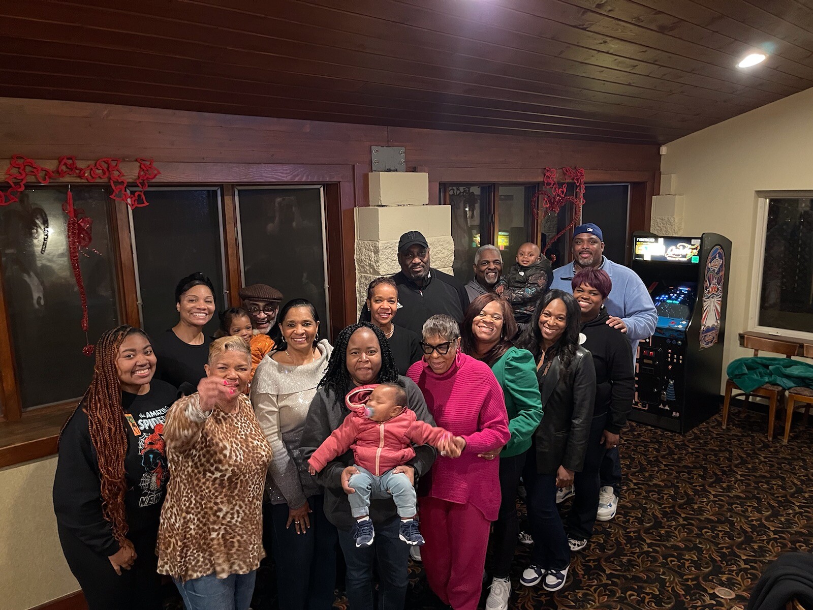 A diverse group of adults and children posing together for a group photo in a warmly lit room with red holiday decorations in the background, everyone smiling at the camera.
