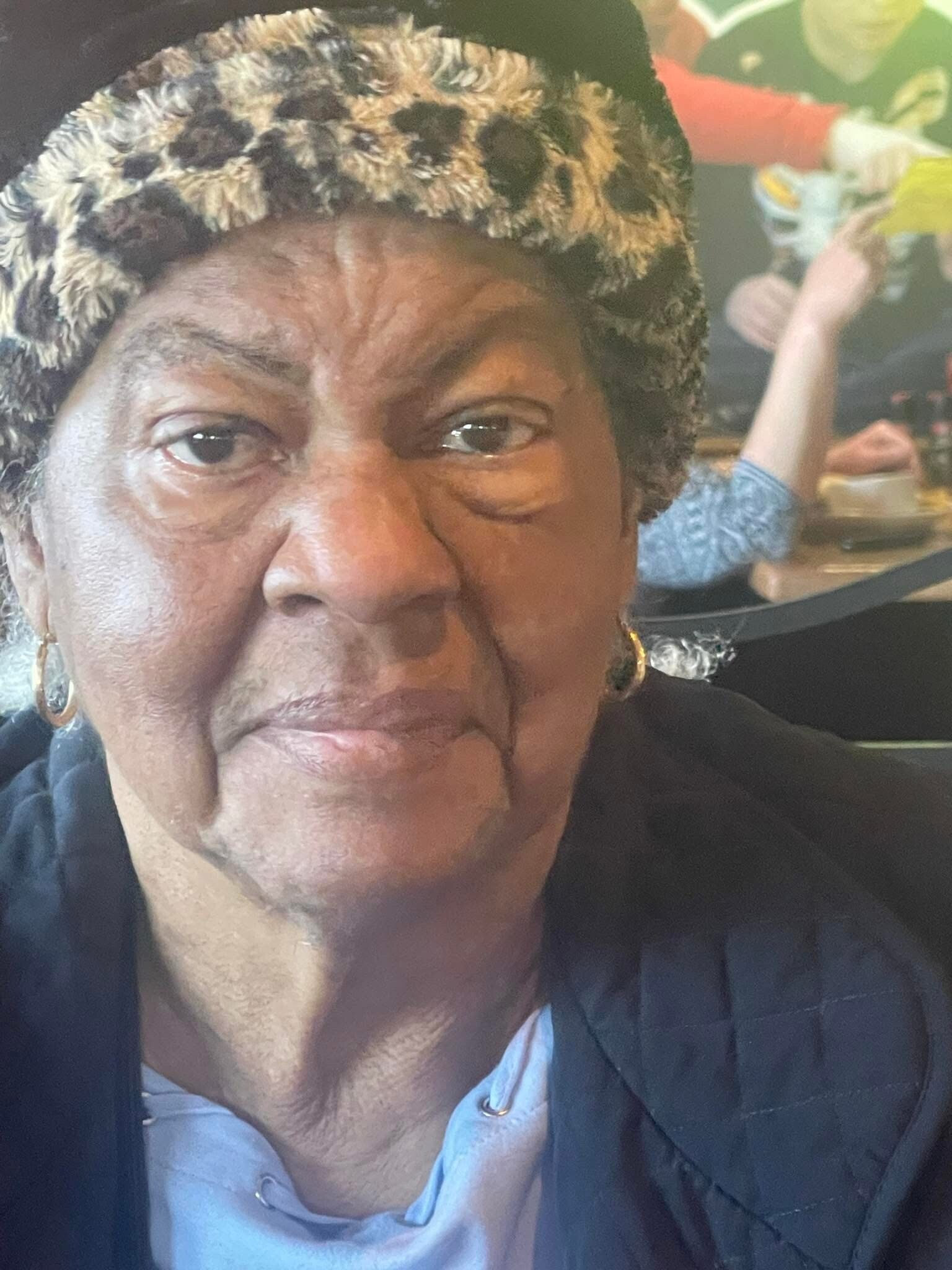Close-up of an older woman wearing a patterned furry hat and gold hoop earrings, with a blurred cafe crowd in the background.