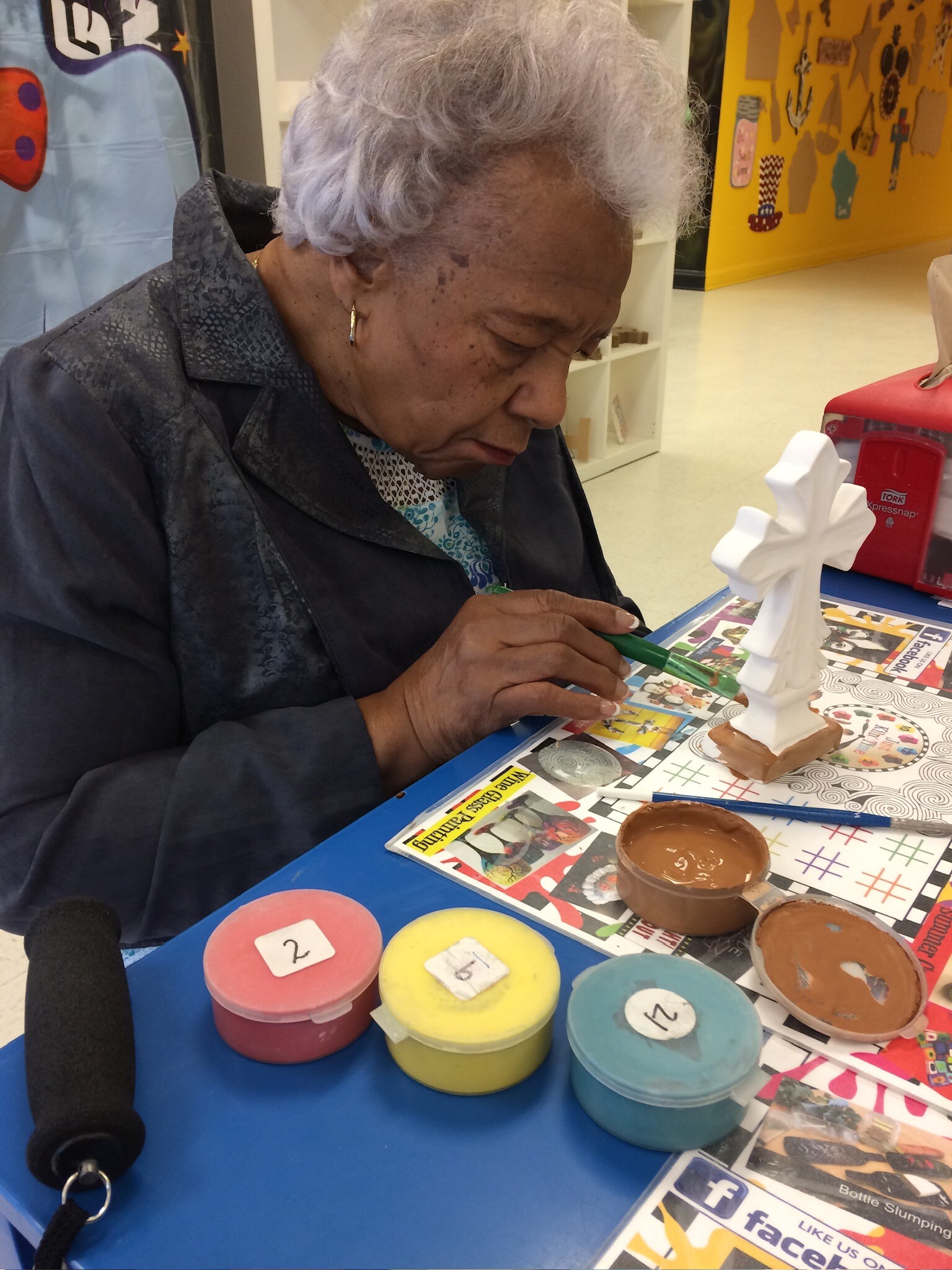 Elderly woman painting a white sculpture at a colorful craft table filled with paint pots in a bright room.