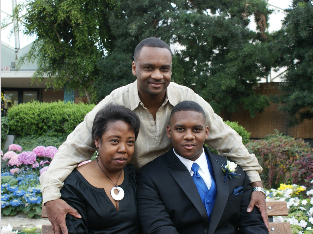 Family portrait: a man in a suit with a blue tie and boutonniere sits with a woman in a black dress as another man stands behind them in a garden.