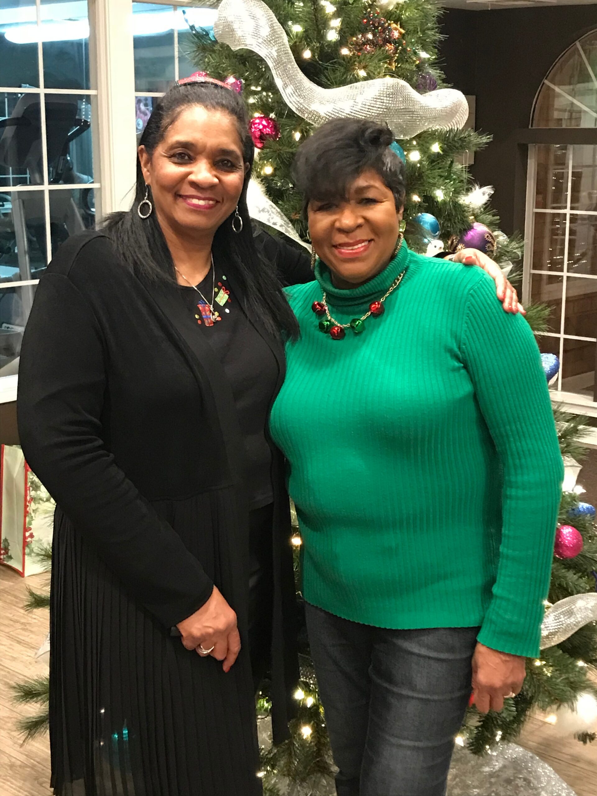 Two women pose together in front of a Christmas tree with lights and decorations, smiling at the camera.