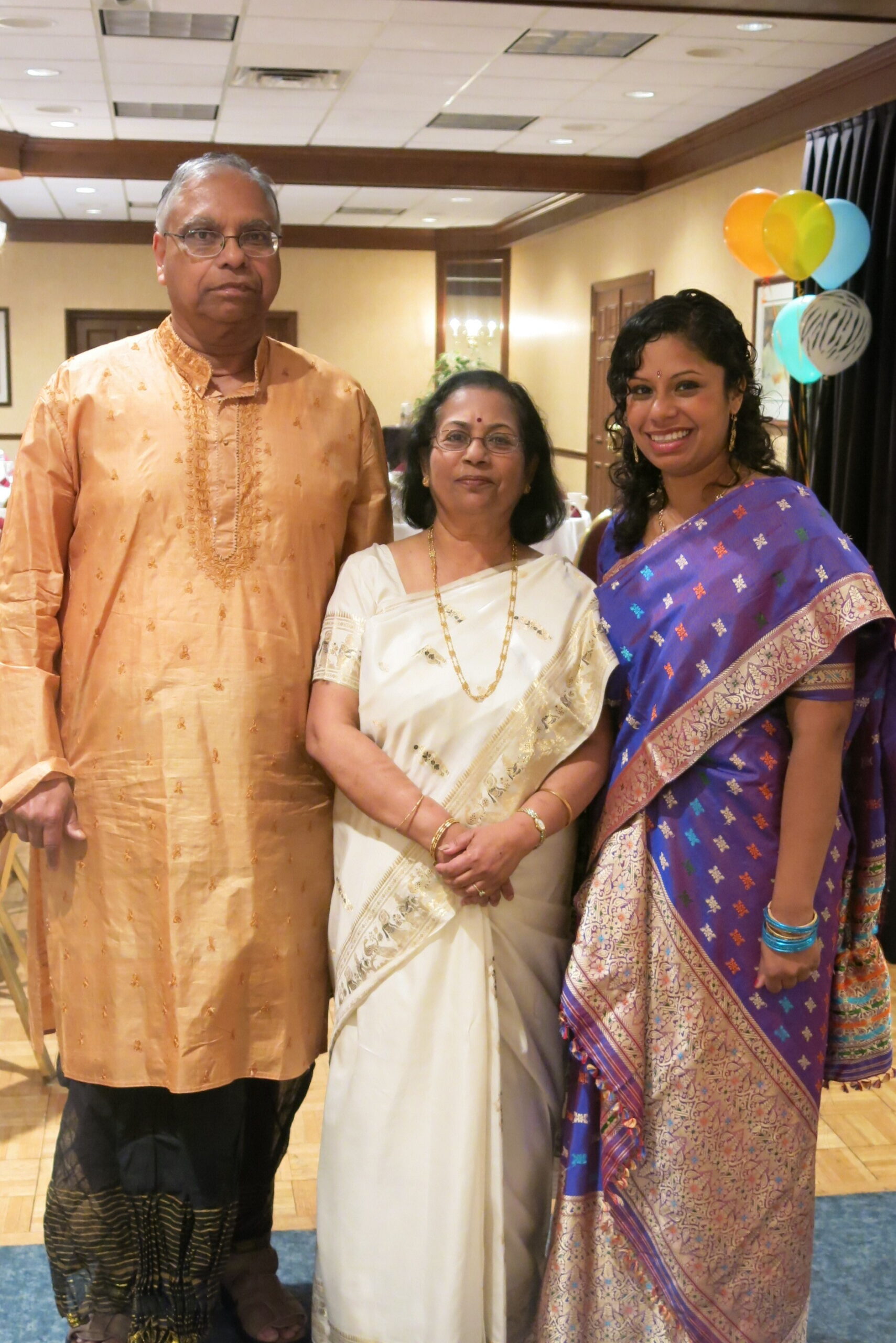 Three adults in colorful Indian attire pose for a celebration photo with balloons in the background.