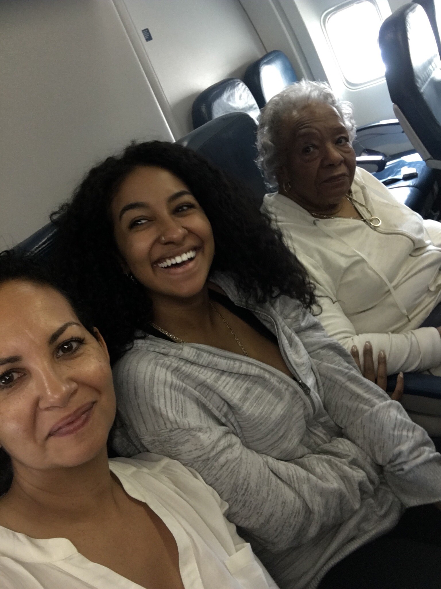 Three women sit close together in an airplane cabin, smiling for a selfie beside the window and seats ahead of them.