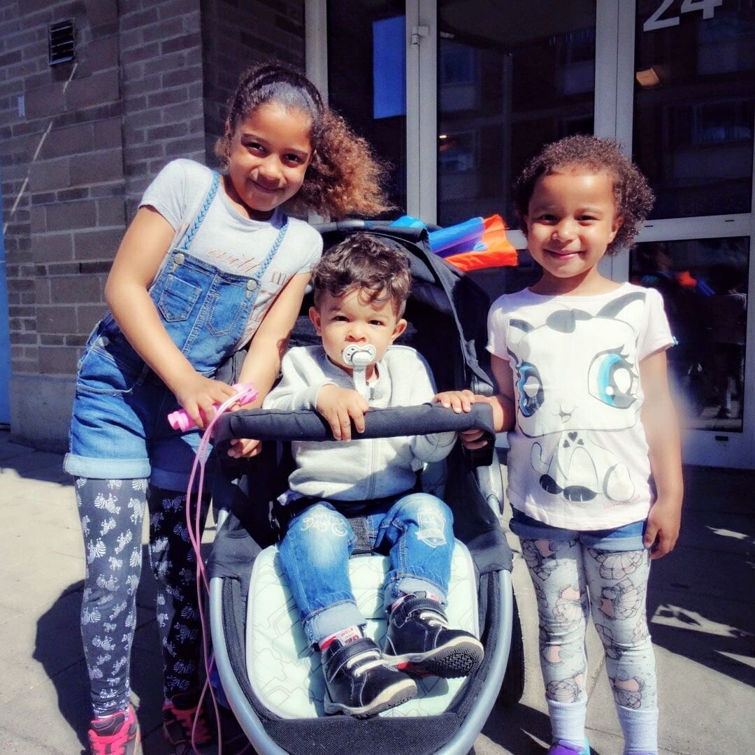 Three smiling children pose together outside a brick building, with a toddler in a stroller between two older girls.