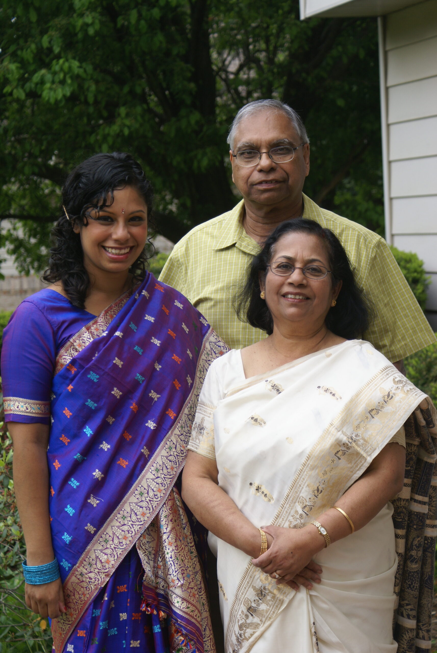 Three smiling people posing outdoors: a young woman in a blue sari, an older woman in a white saree, and a man in a yellow shirt behind them.