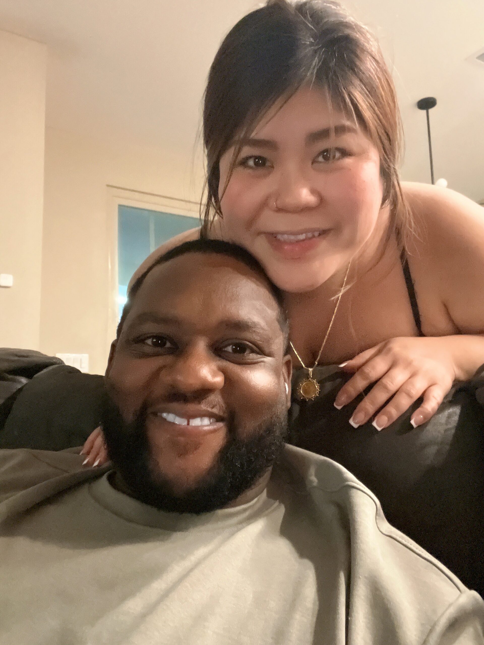 Close-up of a smiling couple posing together, the woman leaning over the man's shoulder in a warm embrace in a cozy indoor setting.