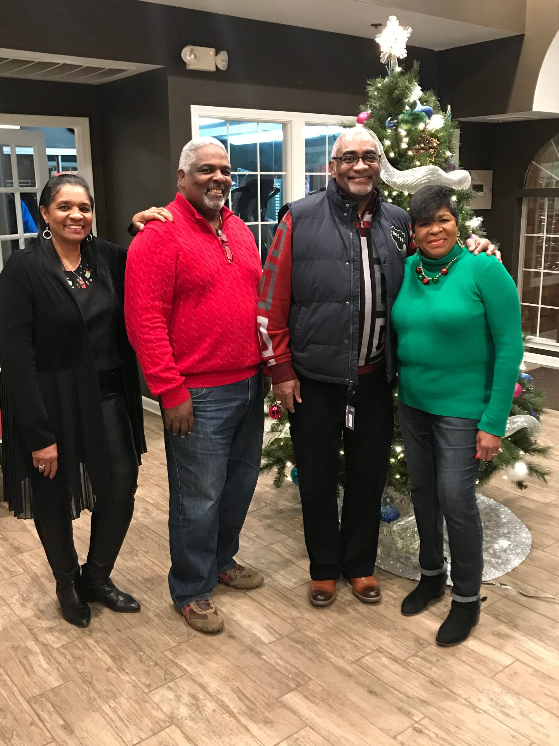Four adults posing together indoors in front of a decorated Christmas tree, smiling for the photo.