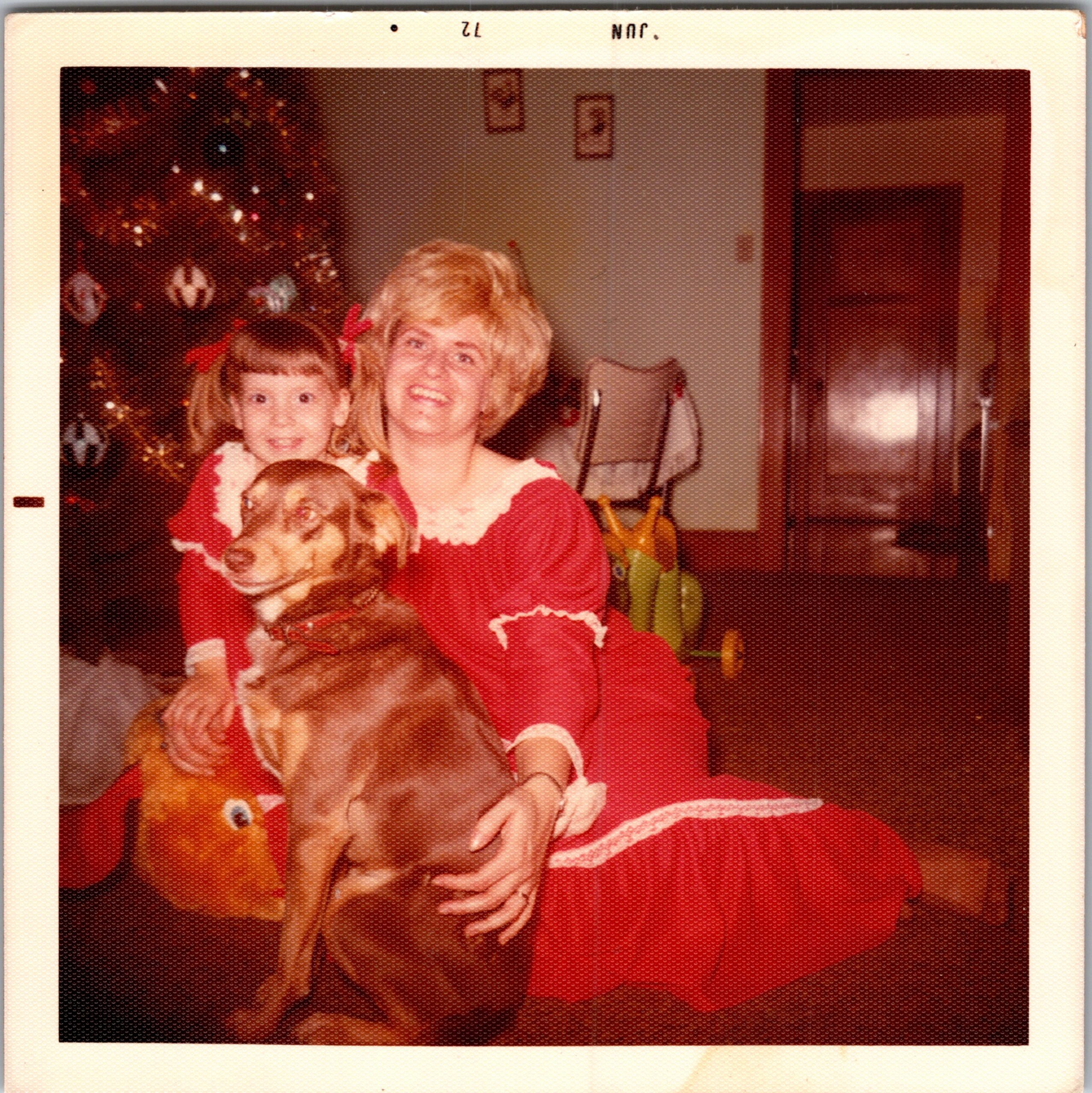 Smiling woman in a red outfit with two children beside her at a Christmas celebration, Christmas tree in the background.