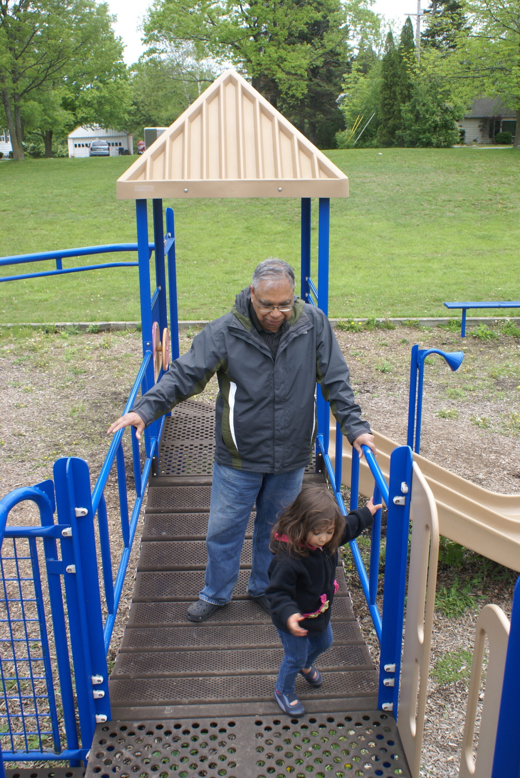 Man guiding a young girl down a perforated metal staircase on a blue playground structure in a park setting.