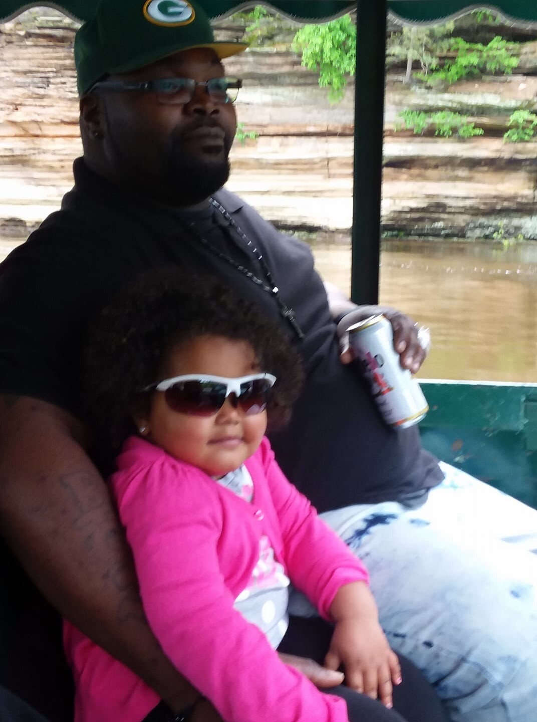 Man and child wearing sunglasses sit together on a boat, the man holding a can of drink, river and rock wall in the background.