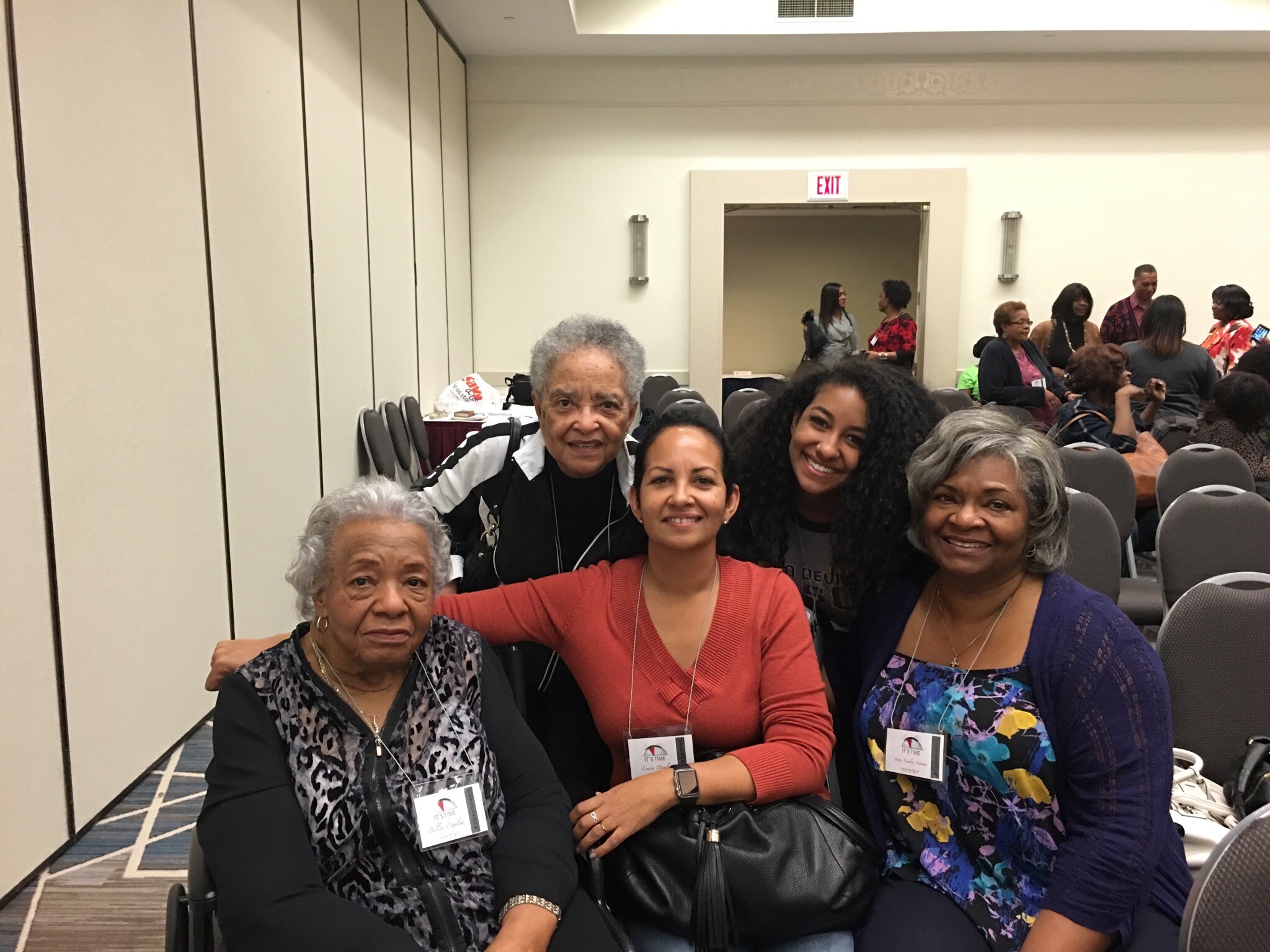 Group of smiling women in a conference room taking a selfie, wearing name badges and lanyards.