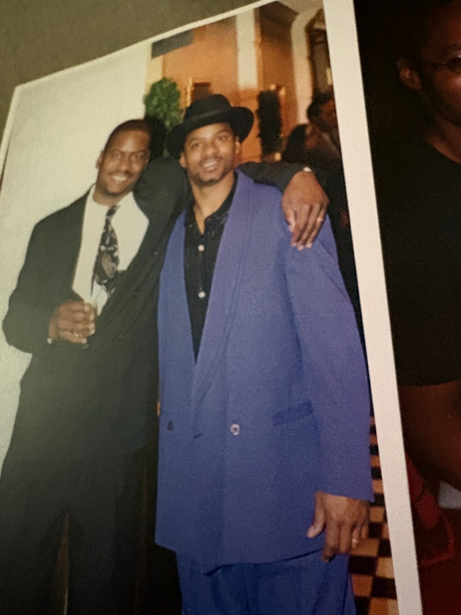 Two men posing for a photo at a party: one in a blue suit with a black hat, the other in a dark suit and patterned tie, sharing a friendly moment.