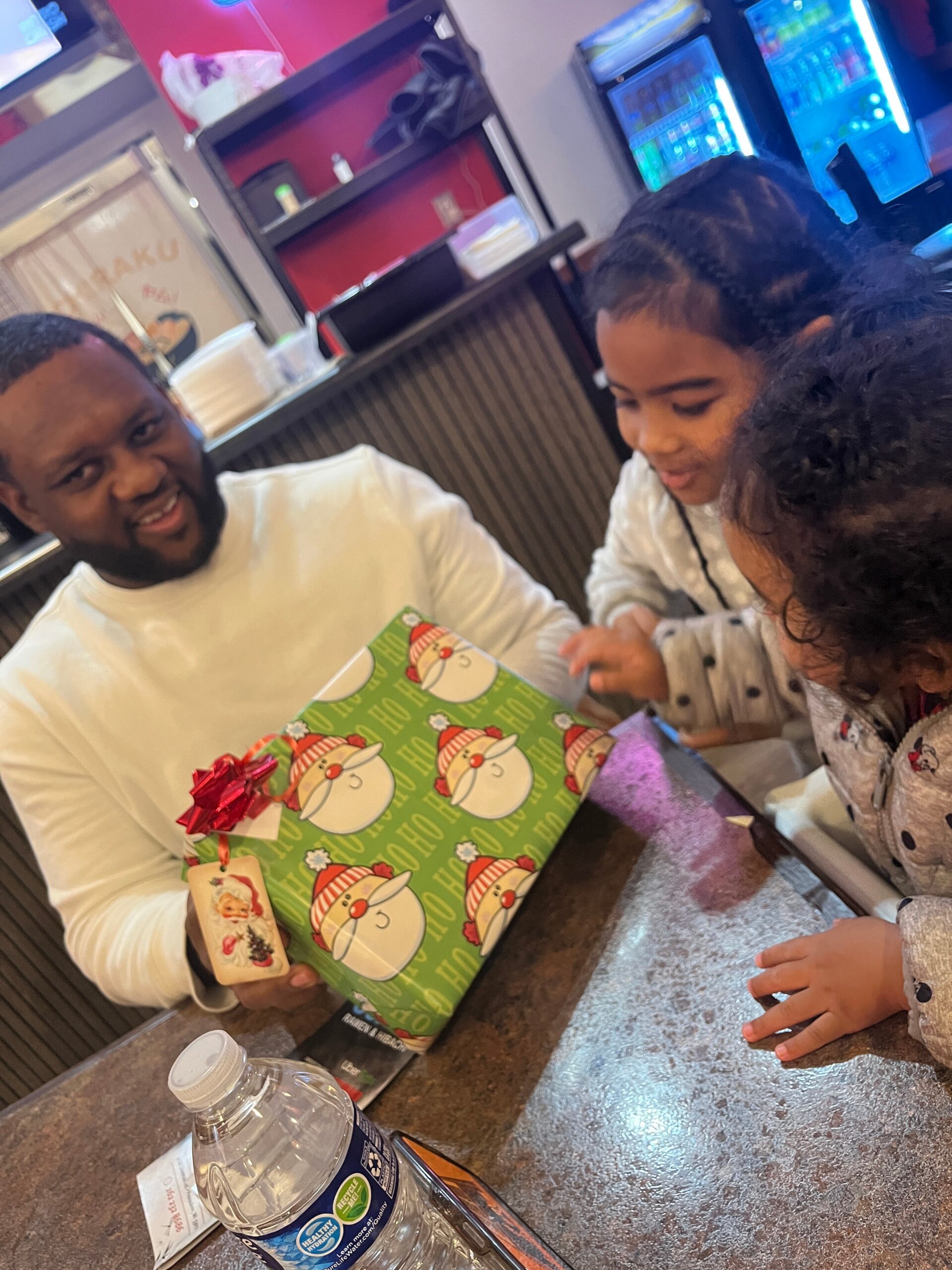Man and young girl sit at a counter, smiling as they unwrap or admire a green Christmas gift bag with snowman faces (holiday scene).