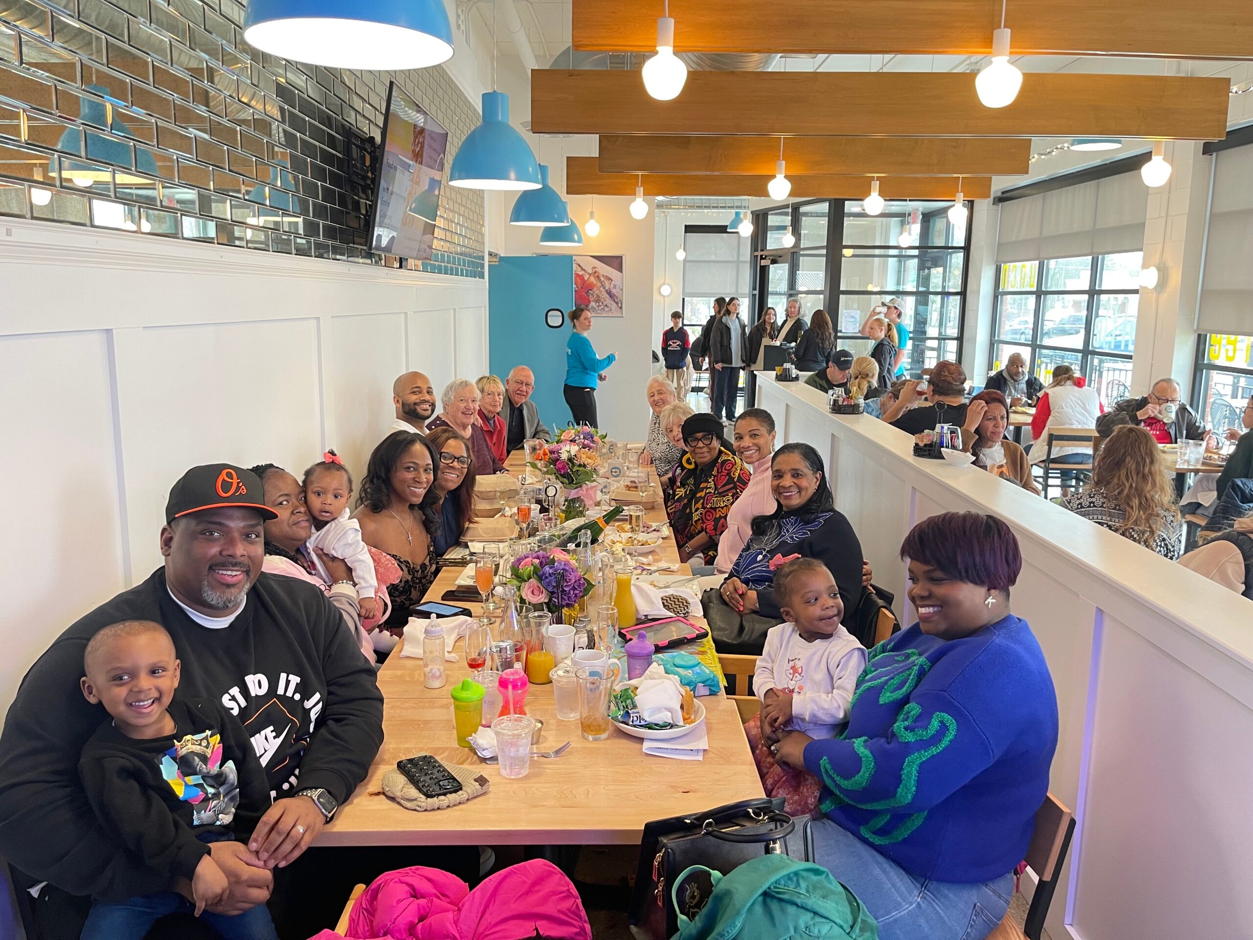 Group of diverse adults and children at a long communal table in a bright cafe, smiling for the camera during a meal party.