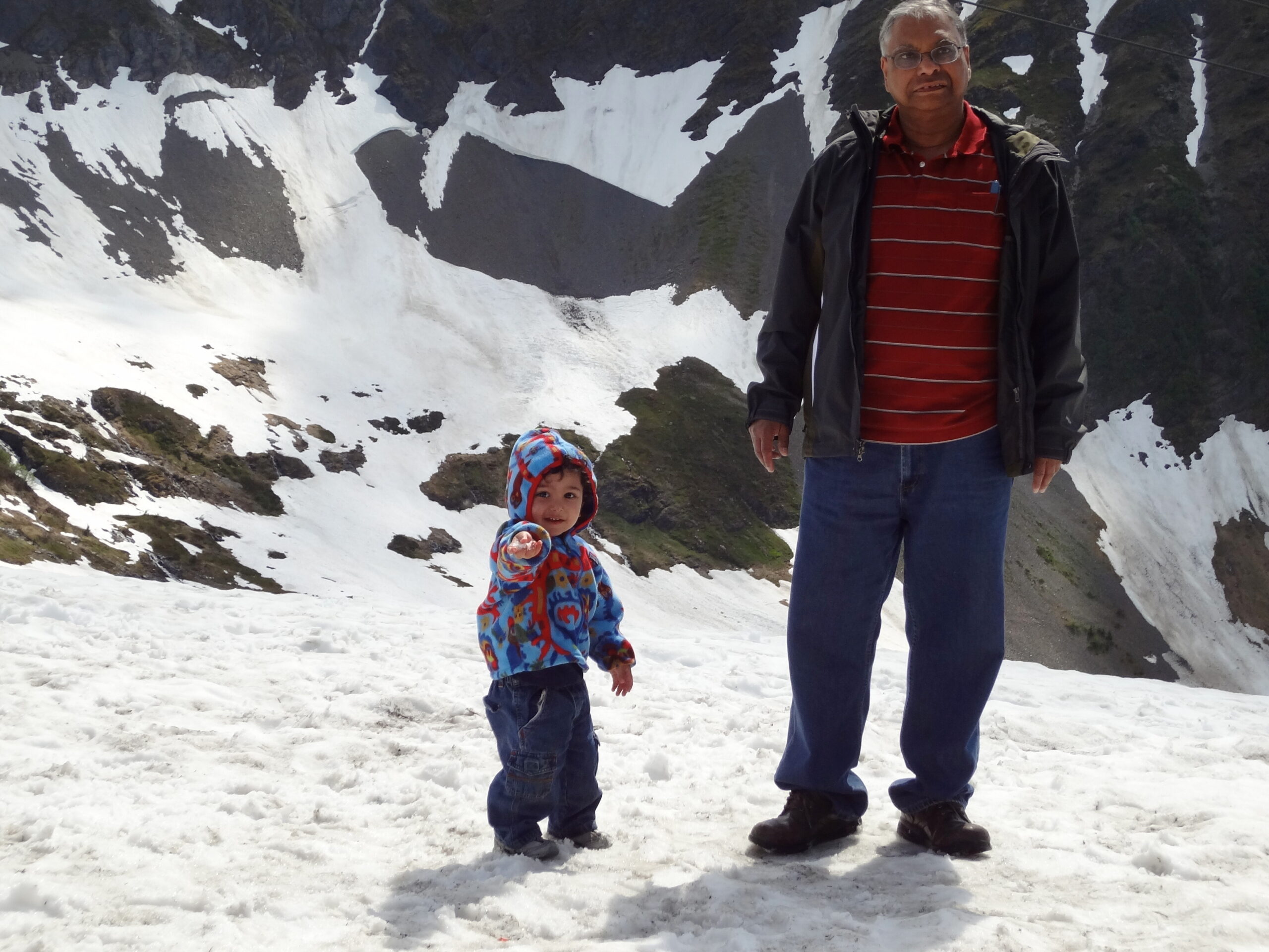 Man in a red striped shirt and jacket standing on snow beside a child in a colorful hooded jacket with a snowy mountain backdrop.