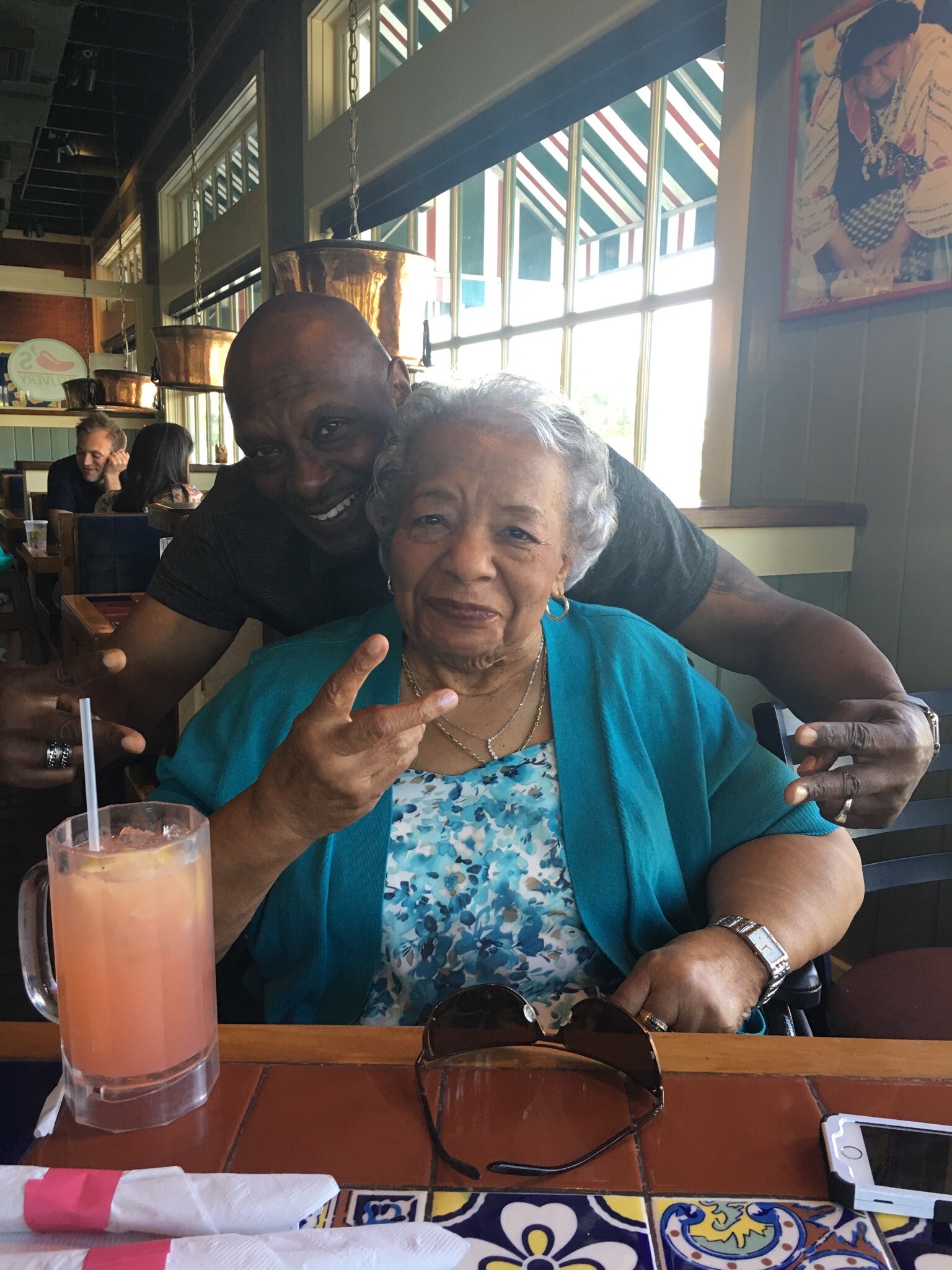 Man and elderly woman smiling at a restaurant table, posing with peace signs; pink drink in foreground and sunglasses on the table.