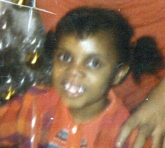 Young child with short dark hair smiling at the camera, wearing an orange shirt at a crowded event.