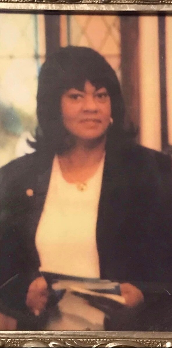 Portrait of a woman with short black hair wearing a dark blazer and white blouse, holding a book or notebook.