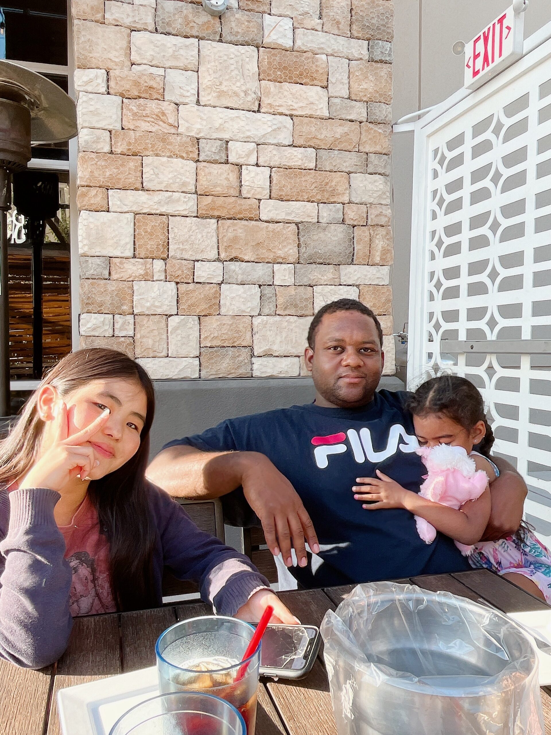 A family posing at an outdoor table: girl peace-signs at the camera, man holding a toddler and baby girl in his arms, drinks on the table, brick wall and EXIT sign in the background.