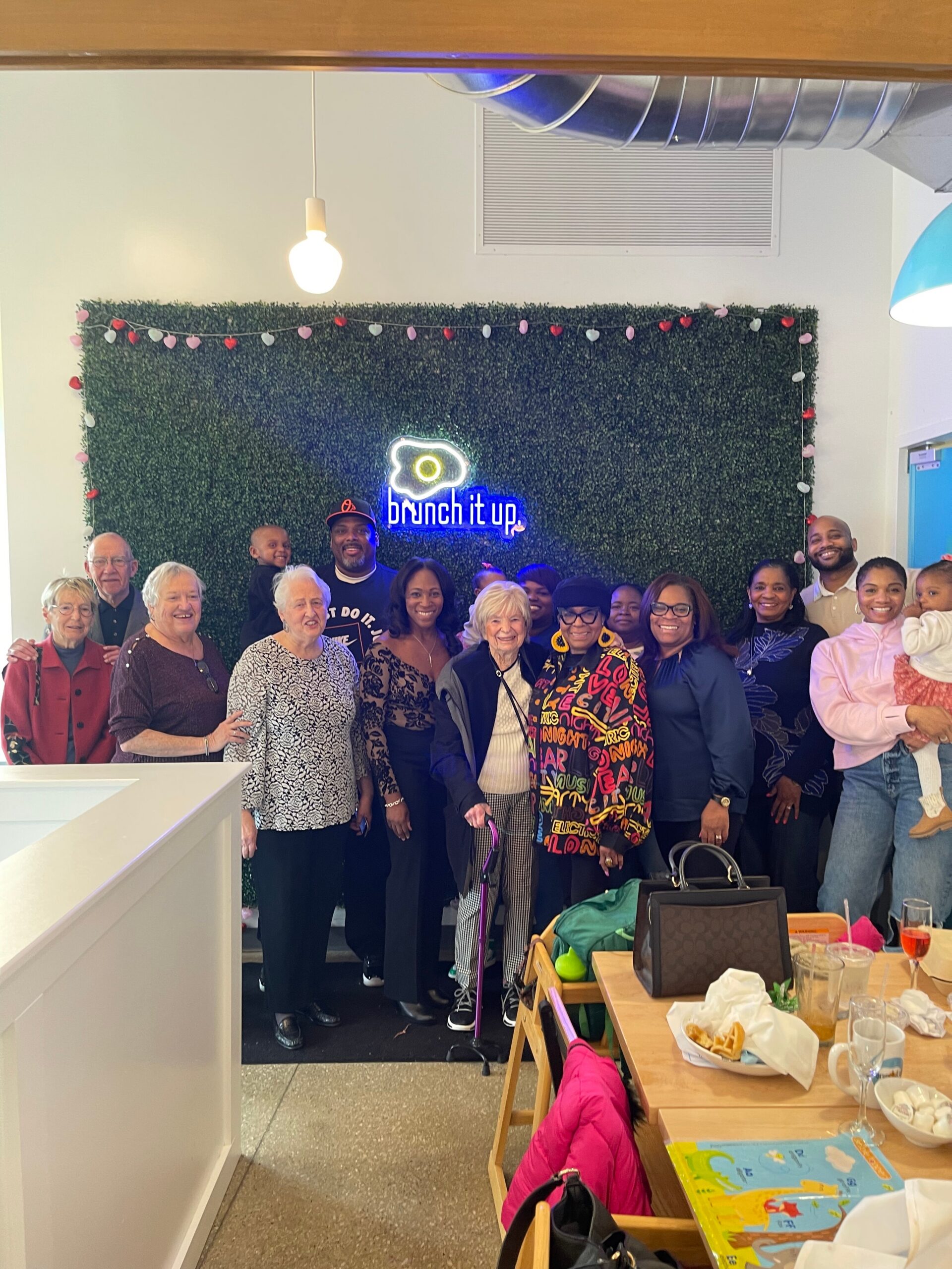 Group of smiling adults and seniors posing in front of a greenery backdrop with a neon 'brunch it up' sign and heart garland at a brunch event, indoors.