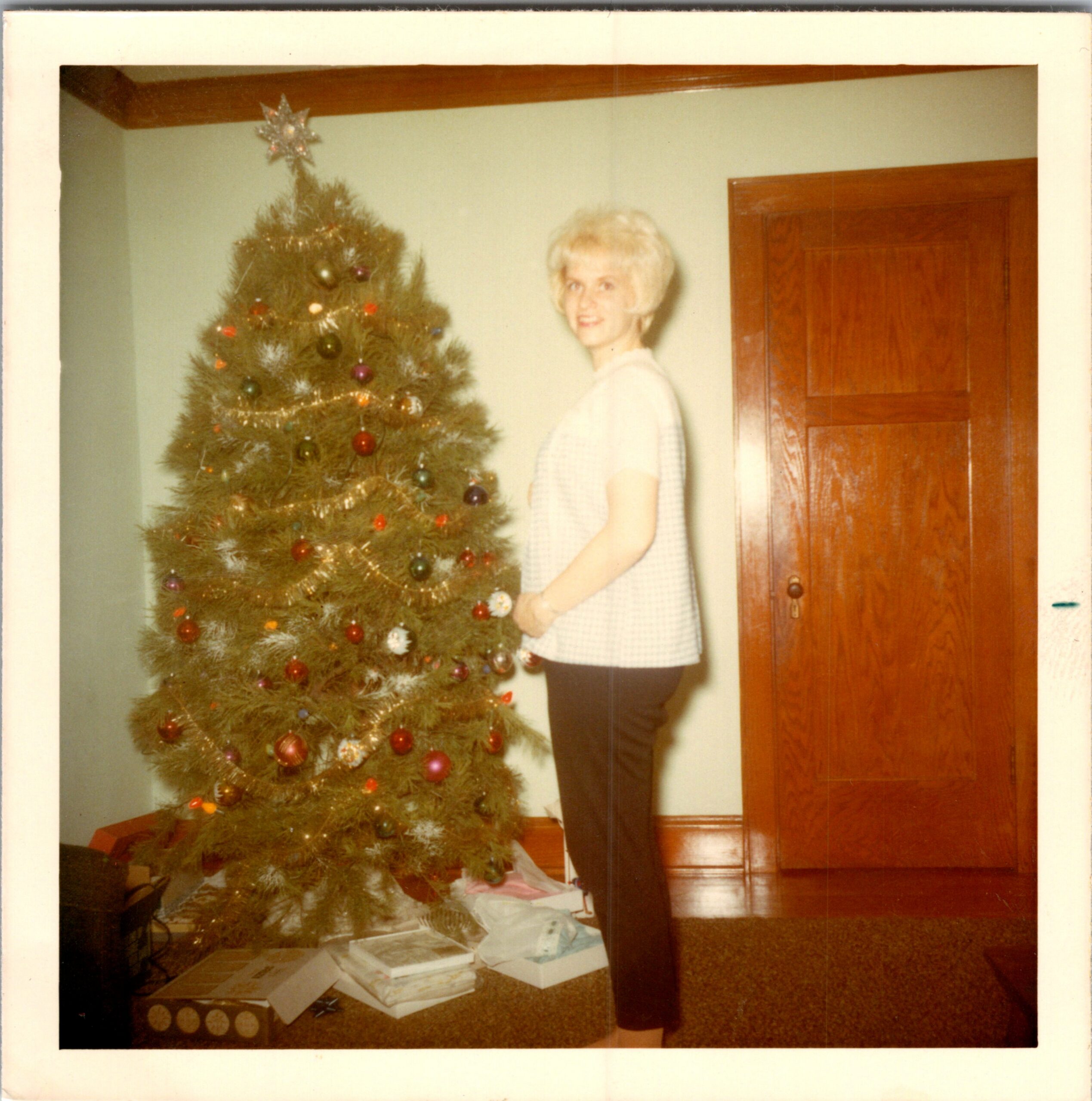 A person with short blonde hair wearing a white sweater stands beside a decorated Christmas tree indoors, with wrapped gifts on the floor nearby.