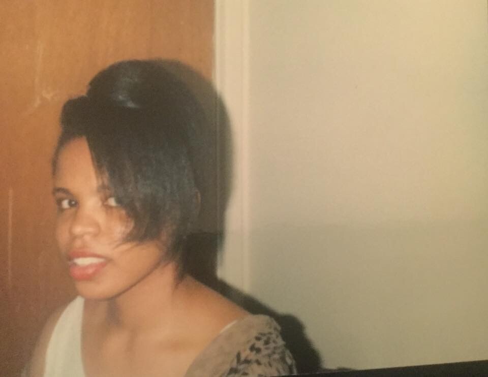 Portrait of a young woman with short black hair, wearing a white top, standing indoors beside a wooden door with a light-colored wall, and a slight smile.