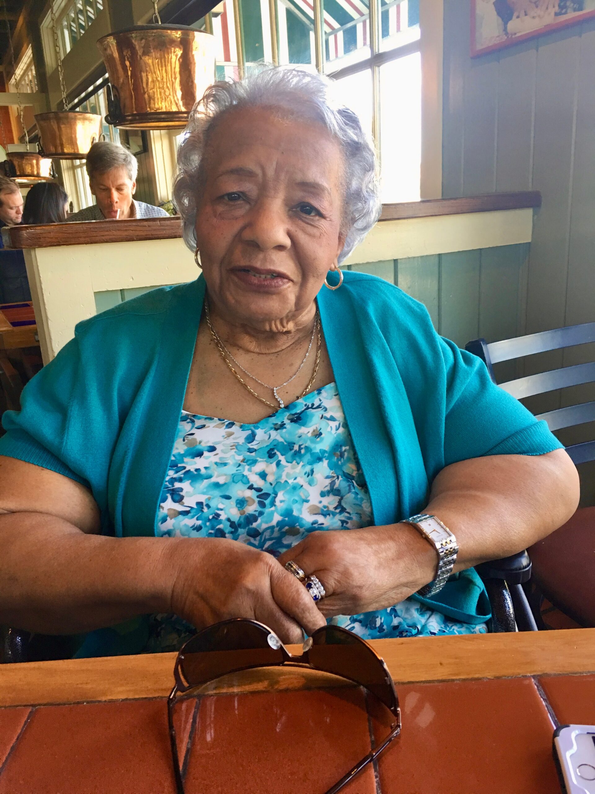 Elderly woman in a turquoise cardigan seated at a cafe table with sunglasses on the table in front and copper pots in the background.